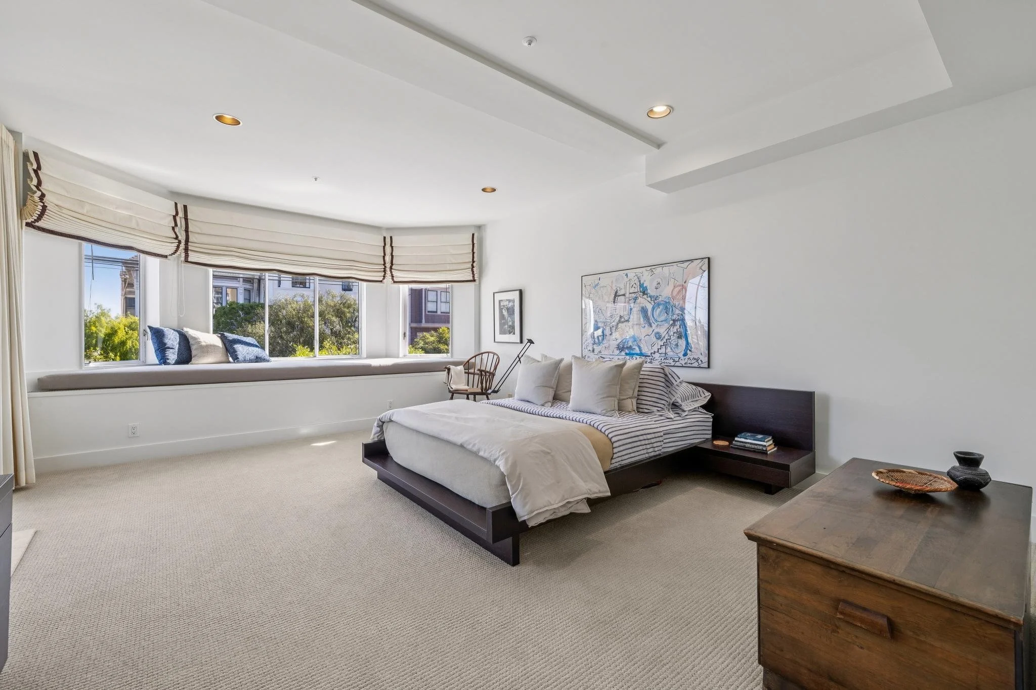 A spacious bedroom with a large bed, a window seat with cushions by a big window, and minimalistic decor.