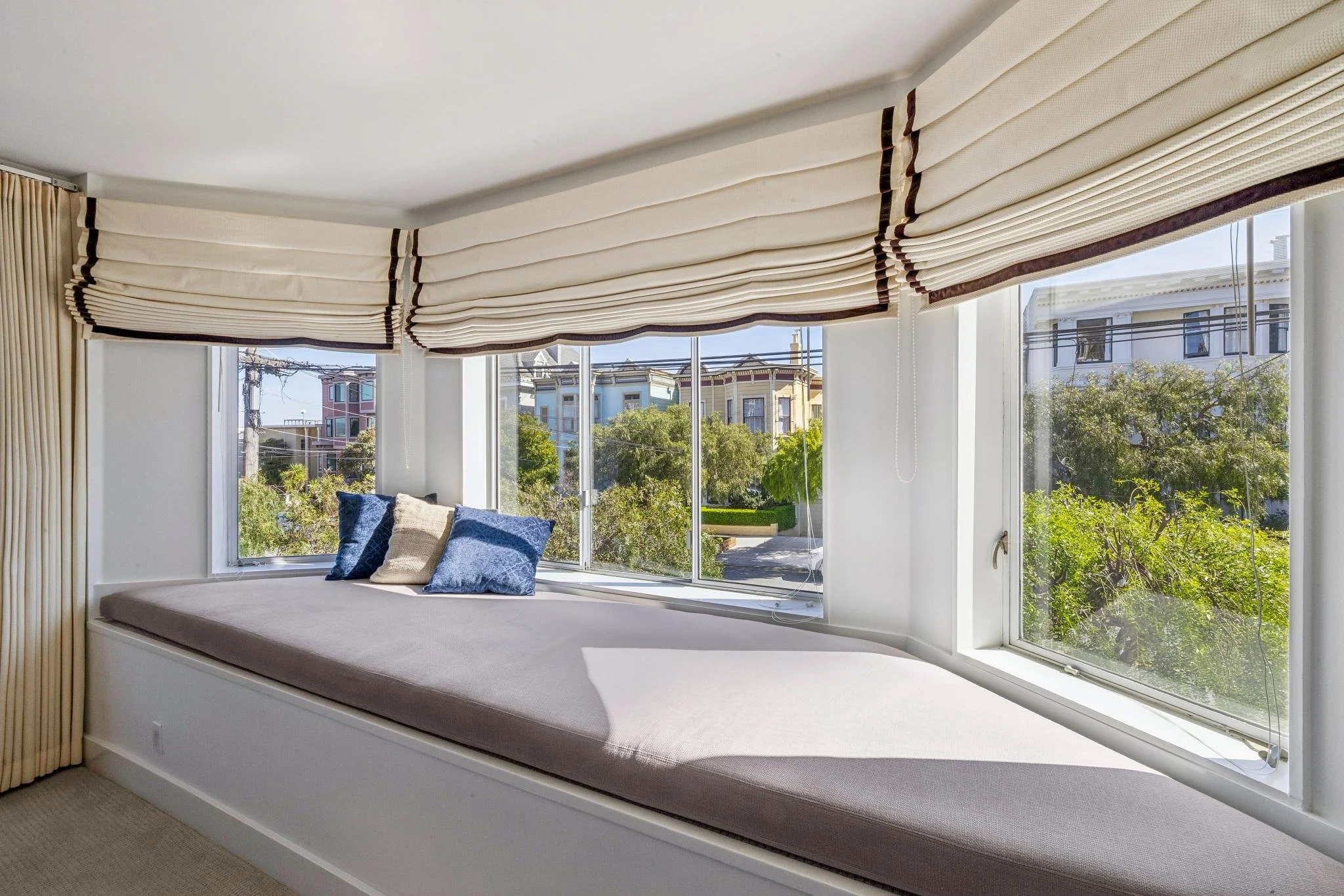 Sunlit window seat with beige cushions and blue pillows, overlooking a tree-lined street with colorful houses.