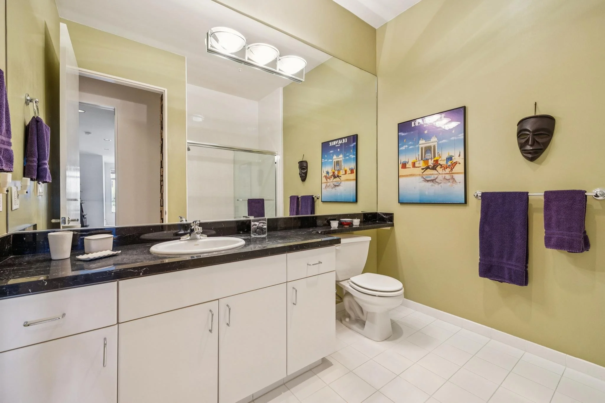 Bathroom with green walls, white cabinets with black countertop, and purple towels on towel rack. A large mirror, wall decor including framed pictures and masks, and a toilet are visible.