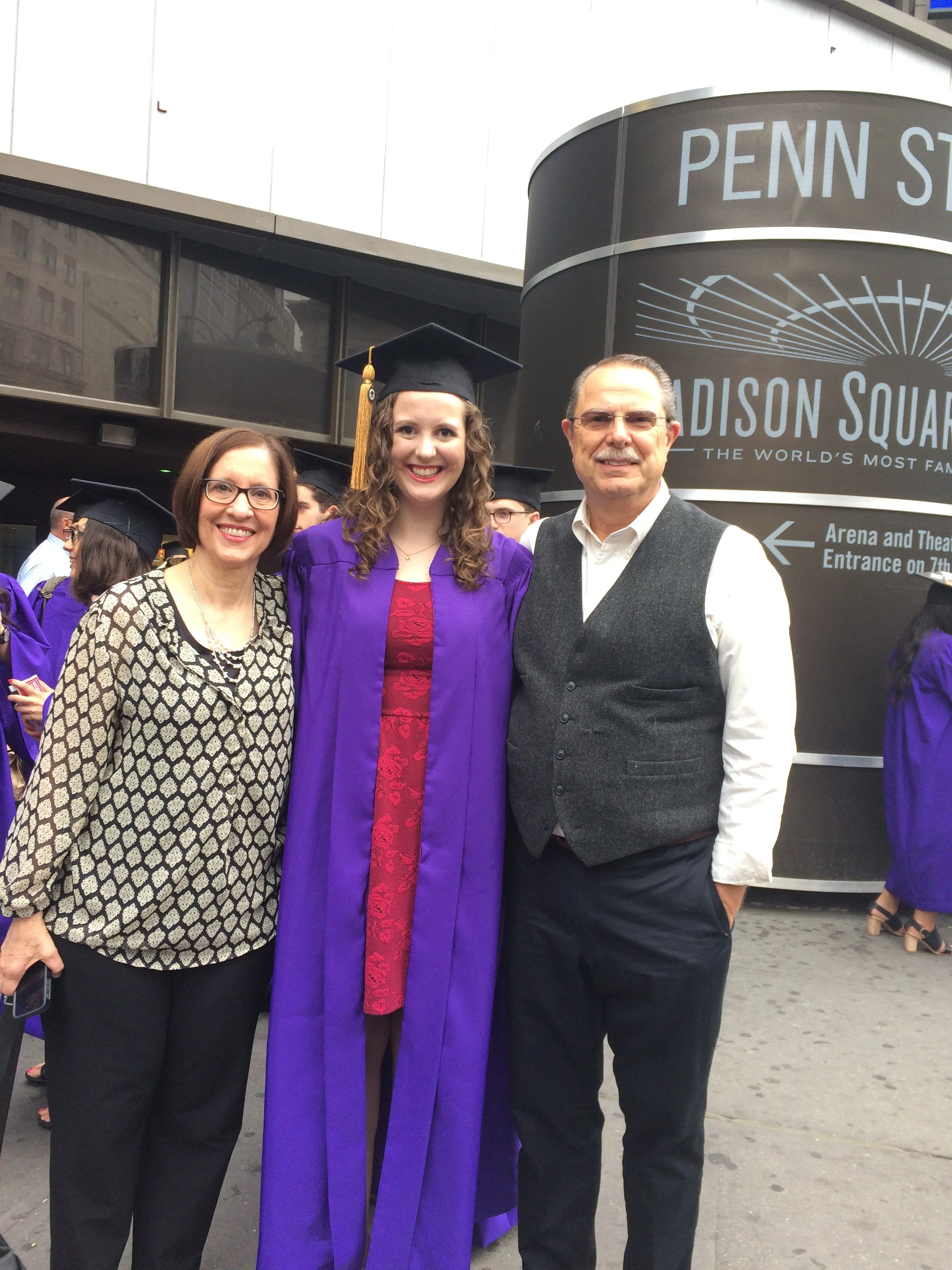 Three people standing outdoors during graduation ceremony, one woman in a purple gown and cap, two others in smart casual attire, with Penn Station sign in the background.