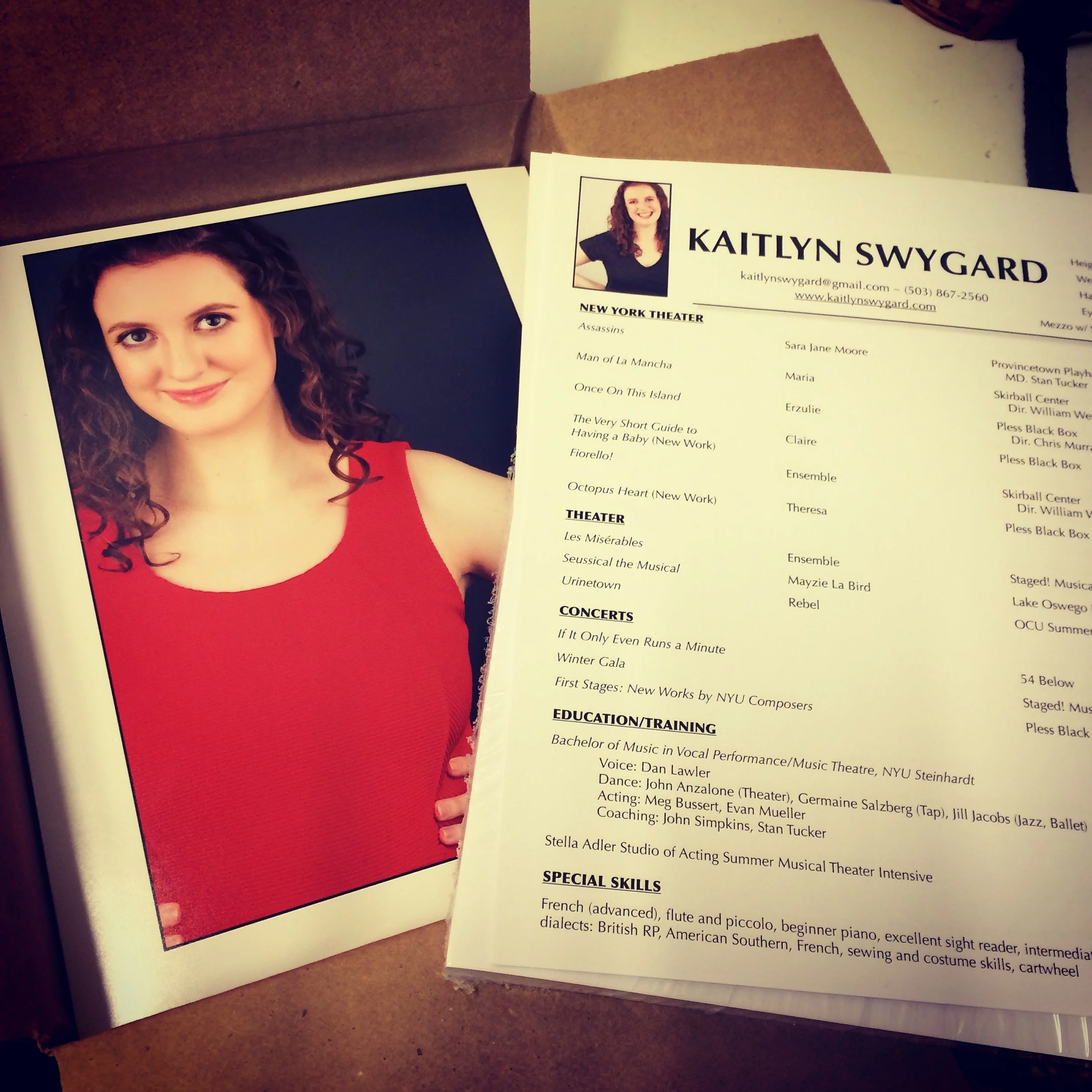 Open booklet inside a person's hand, showing a professional headshot of a woman with curly hair and a lengthy list of theater performances, contact information, and skills. The other page displays a large red shape.