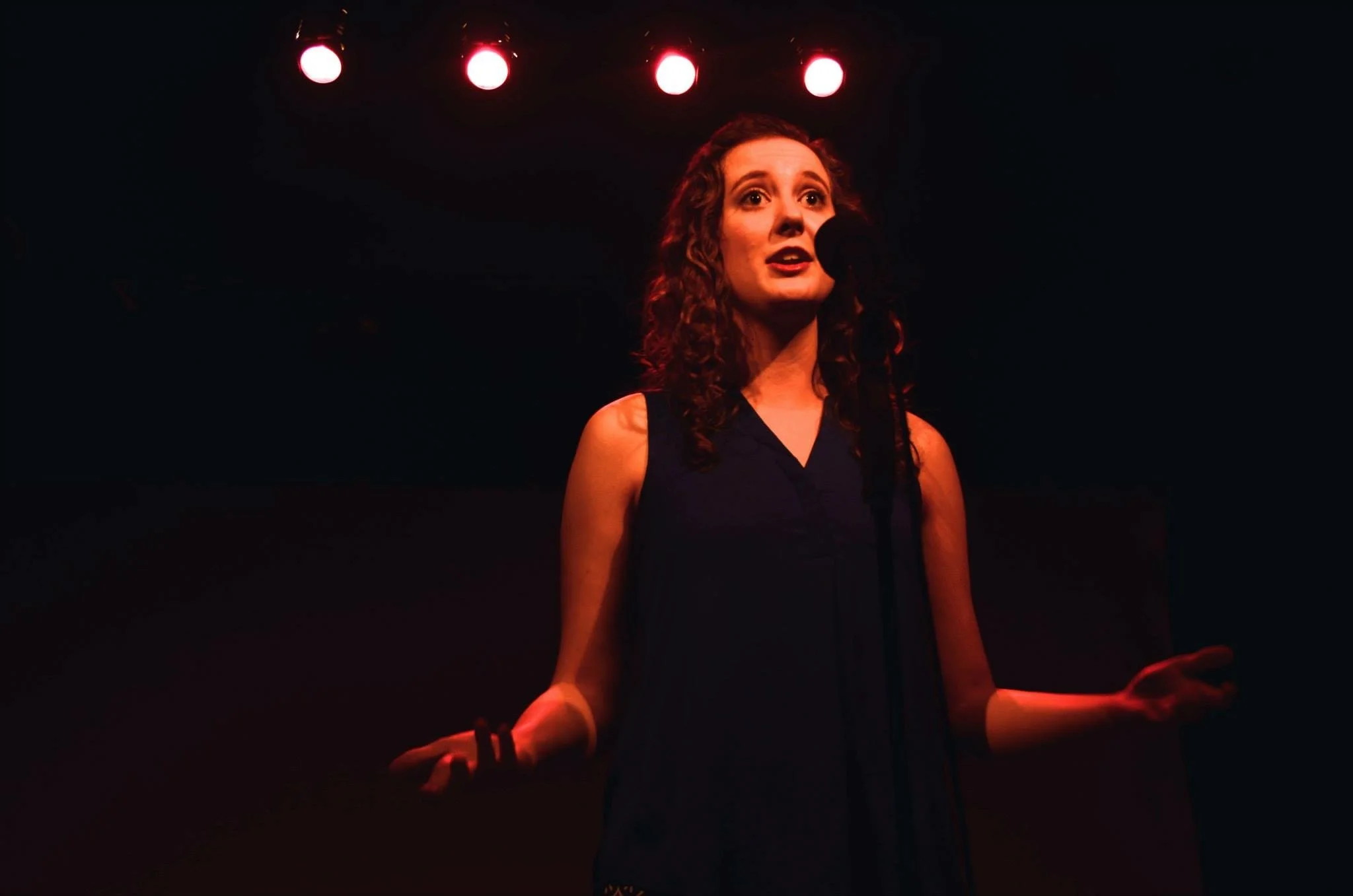 A woman standing in a dark room under red stage lights, speaking or singing into a microphone.