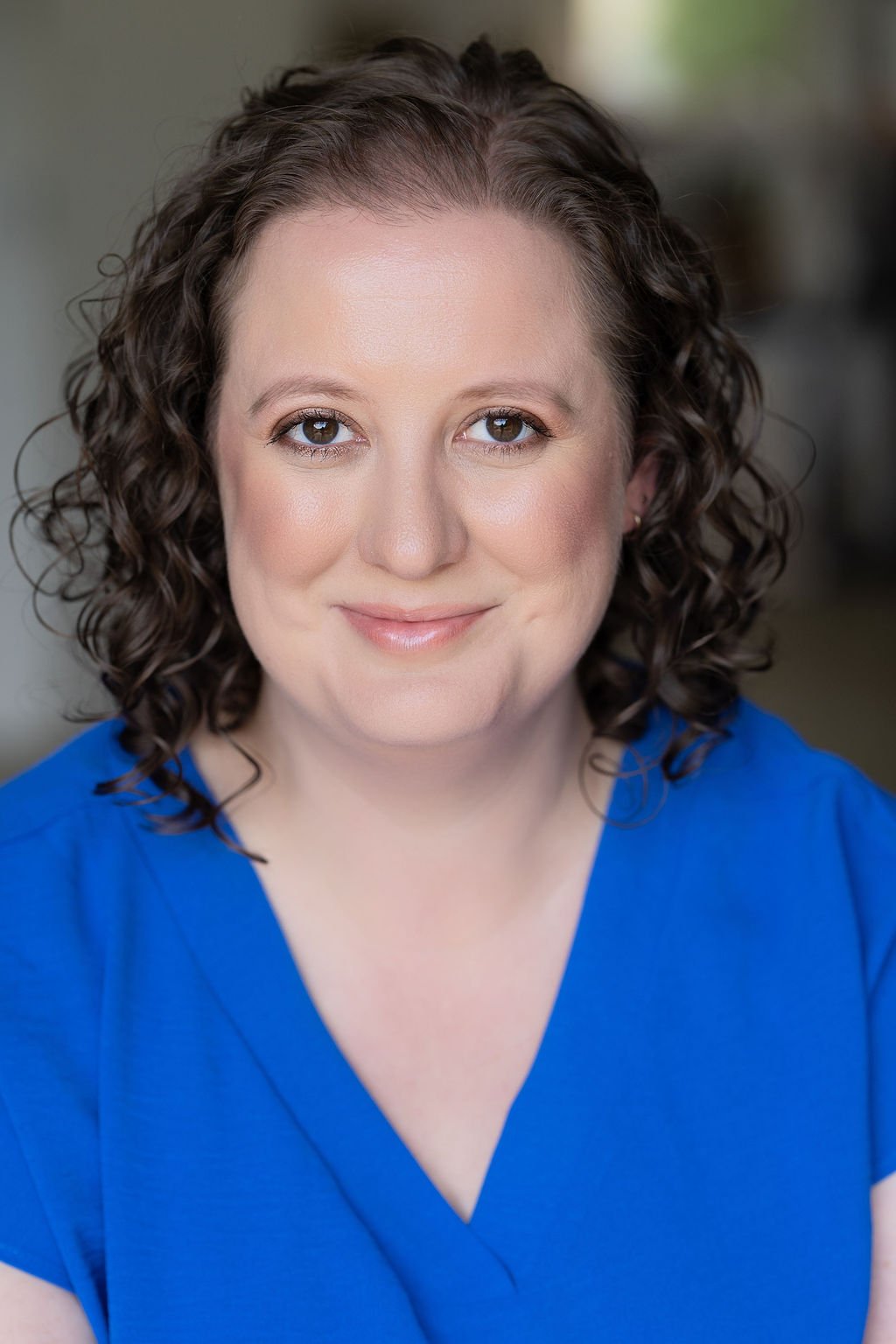 A woman with curly brown hair and brown eyes smiling, wearing a blue top, in a close-up portrait with a blurred neutral background.
