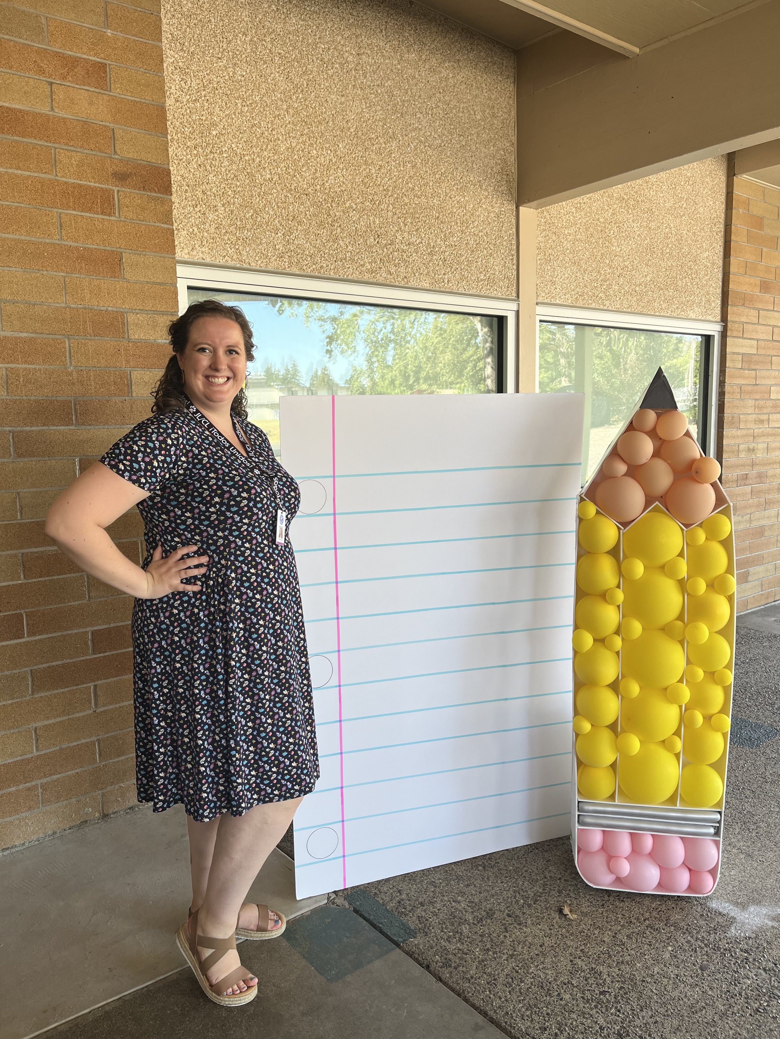 A woman standing outside near a large notepad and a balloon display with pink, yellow, and peach-colored balloons arranged in a pyramid shape.