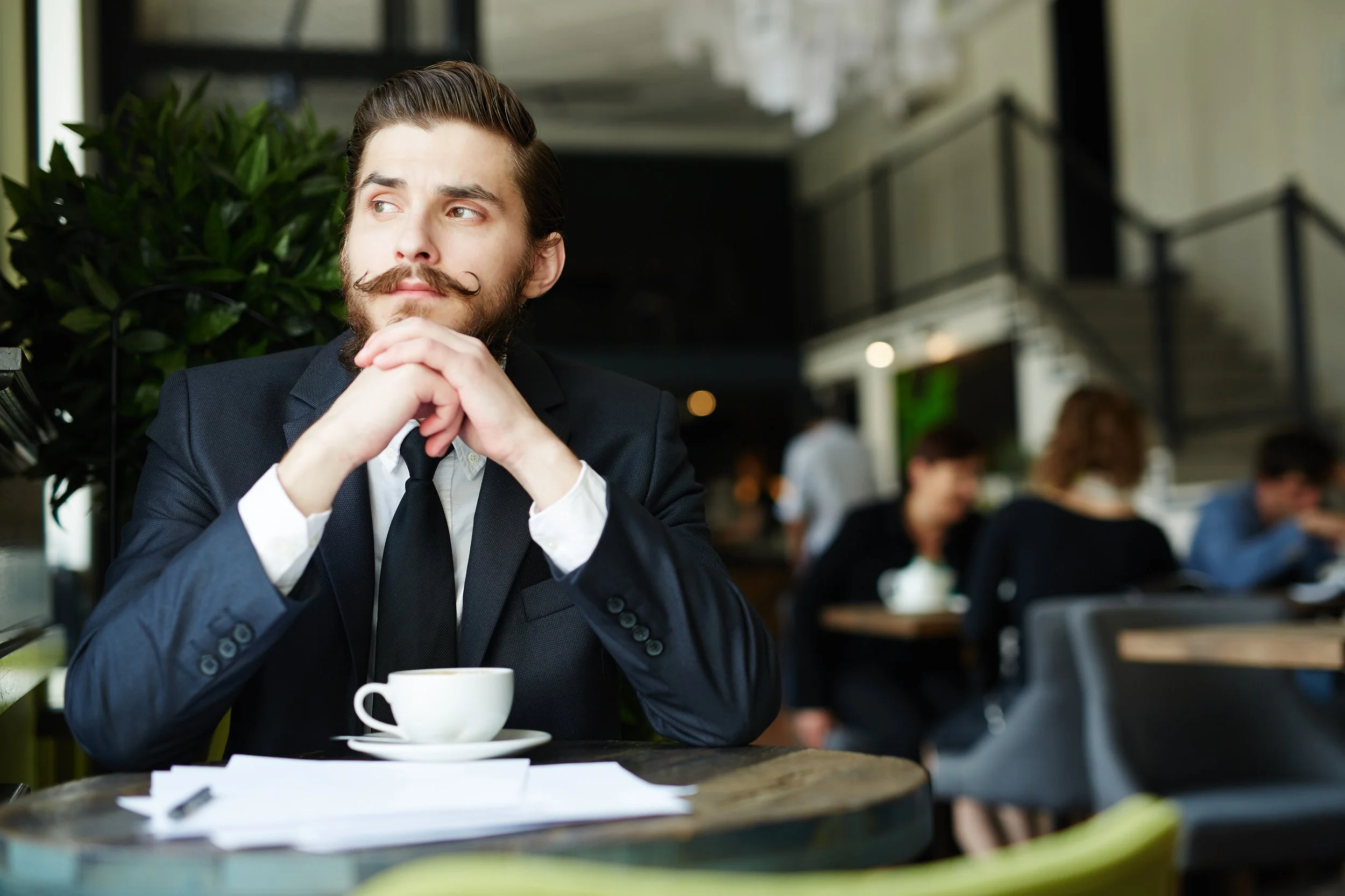 Un homme d'affaires assis dans un café, portant un costume noir, avec une tasse de café devant lui. Il semble pensif, regardant à gauche, avec d'autres clients en arrière-plan.
