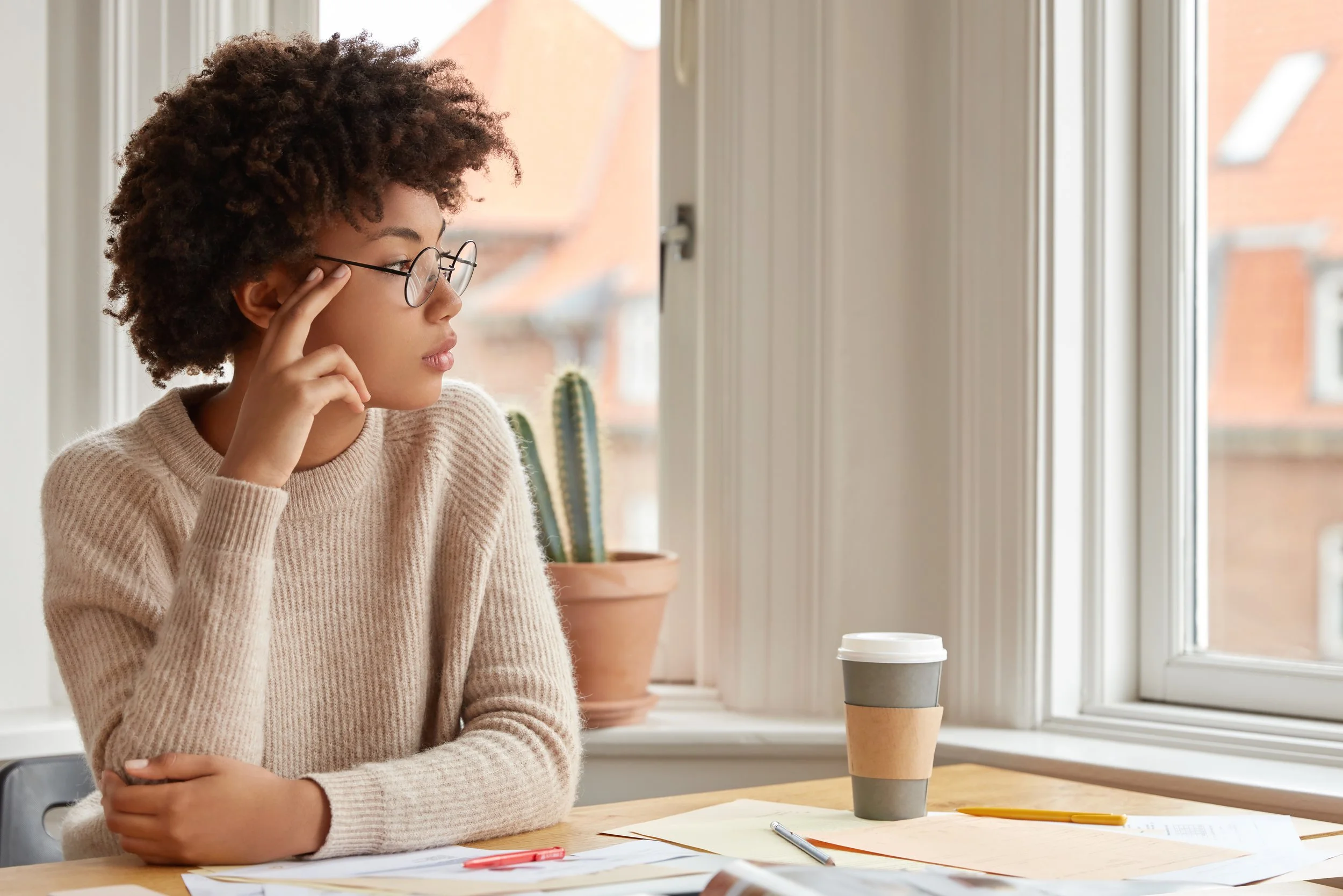 Une femme avec des cheveux bouclés, portant des lunettes, assise à une table près d'une fenêtre avec une tasse à café, des papiers et des stylos.