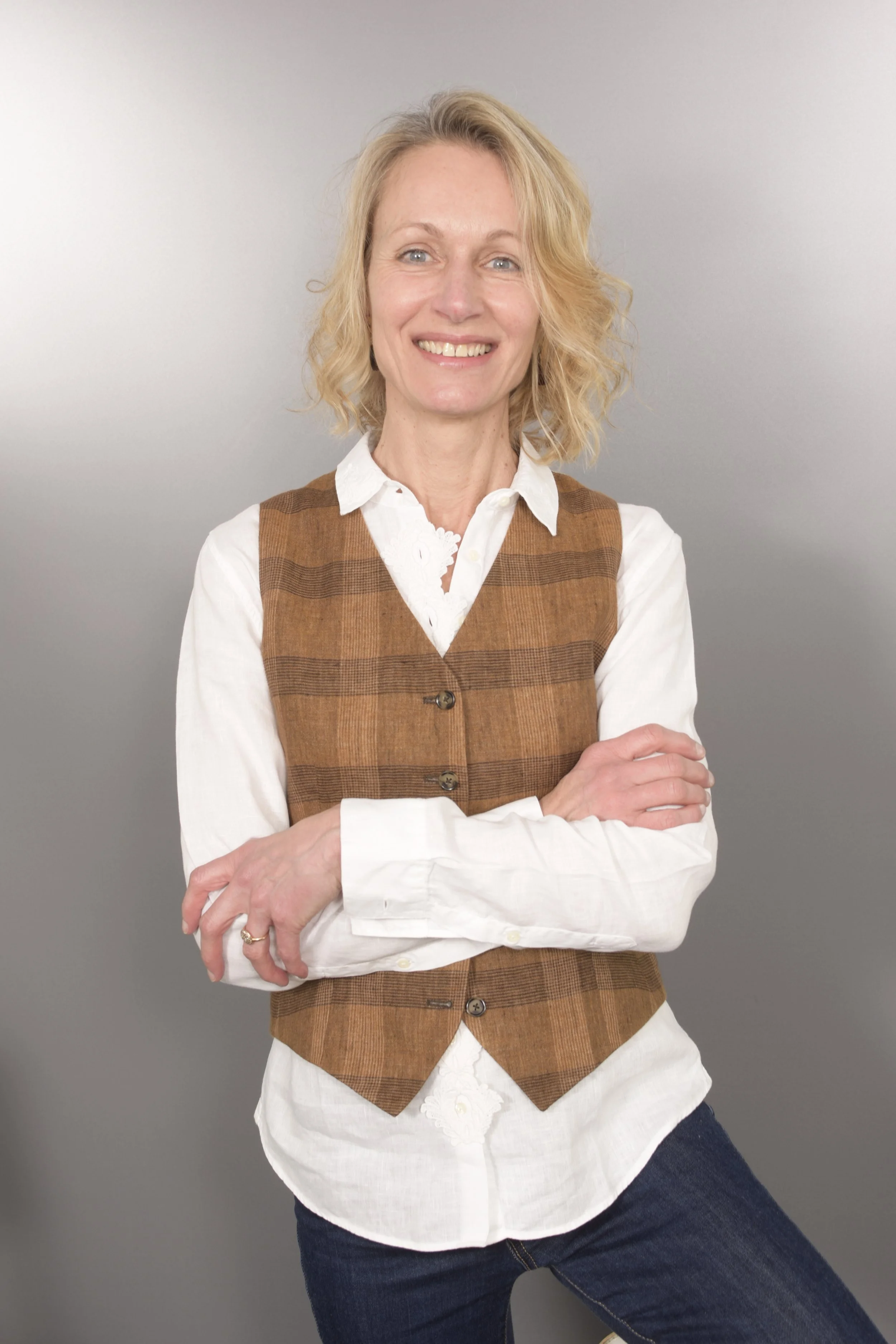 Une femme blonde souriante, aux cheveux bouclés, portant une chemise blanche avec un veston marron à carreaux et un jean foncé, se tenant contre un fond gris clair.