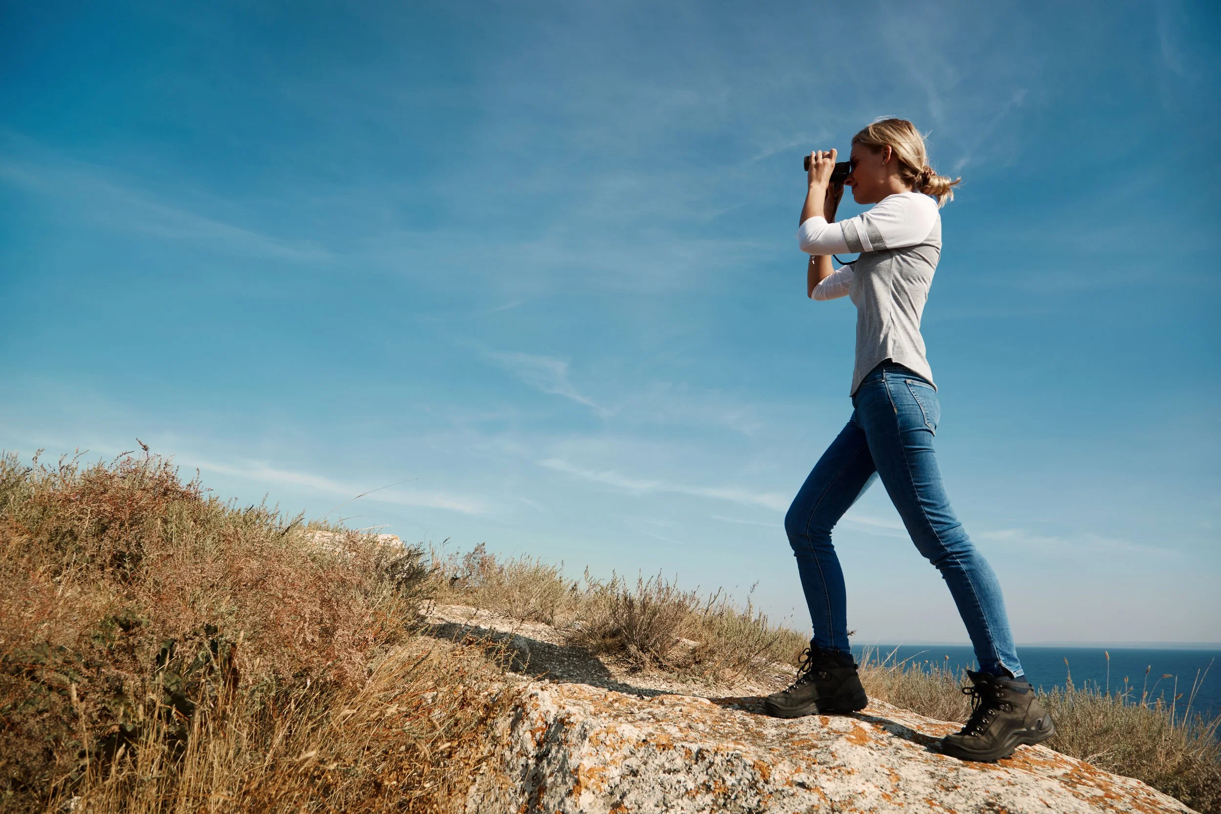 Femme faisant de la randonnée en paysage côtier, regardant à travers des jumelles, portant des chaussures de marche, sous un ciel bleu clair.