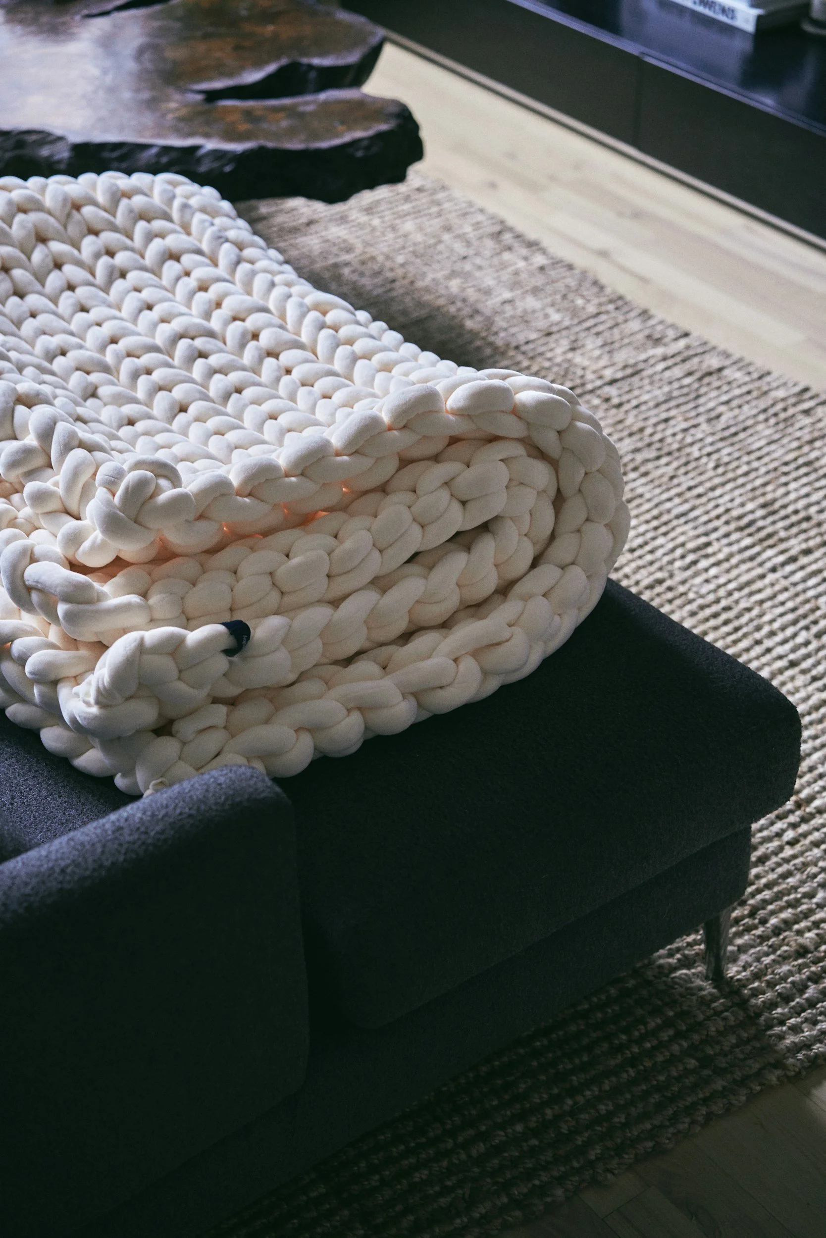Close-up of a chunky white knitted blanket on a black sofa in a modern living room.