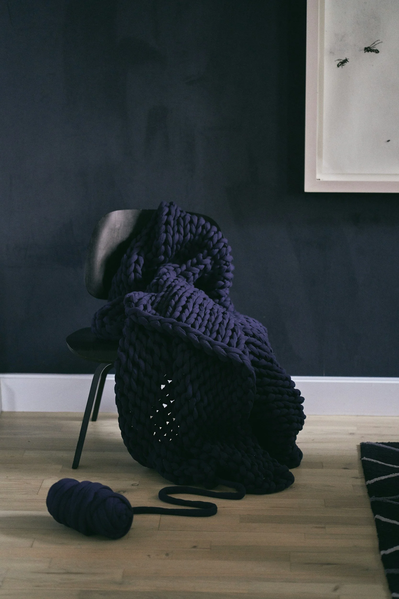 A black chair draped with a large, chunky, dark purple knit blanket, with a matching ball of purple yarn on the wooden floor beside it, against a dark wall with a framed whiteboard on the right.