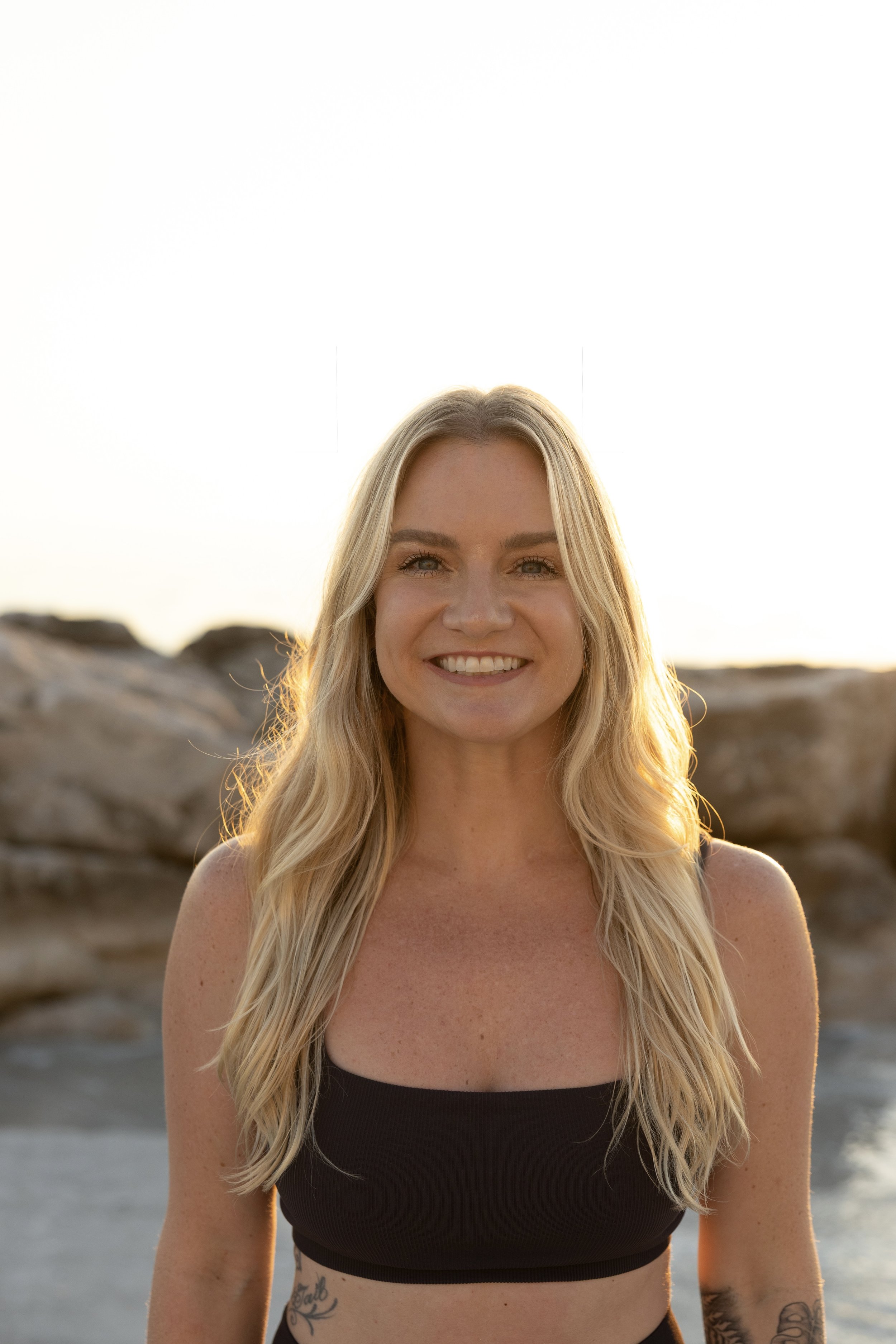 A smiling woman with long blonde hair standing on a beach at sunset, with rocks in the background.