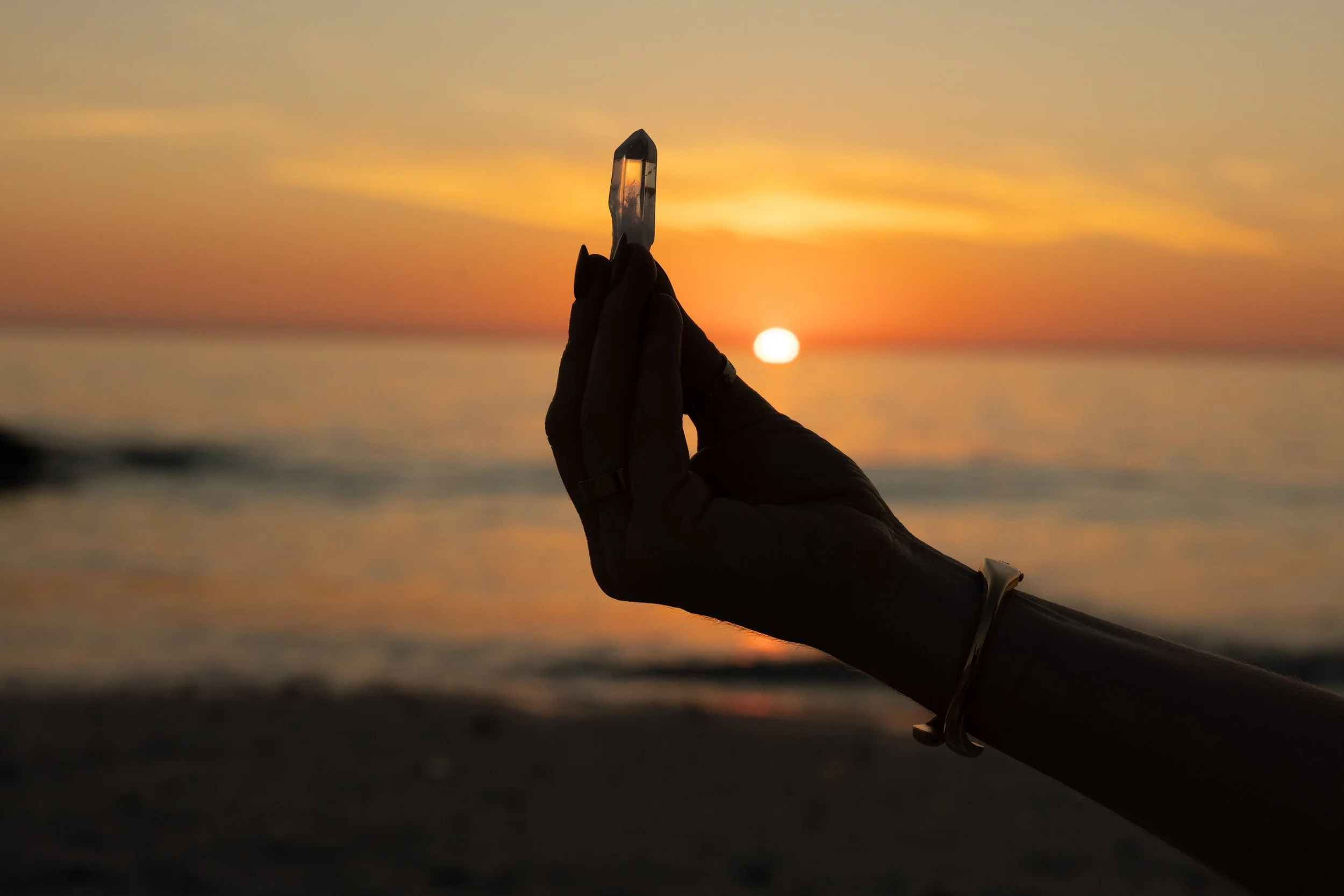 A shadowed hand held out, palm up while holding a clear crystal within the fingers. The background is sunset at the ocean, while standing on the beach.