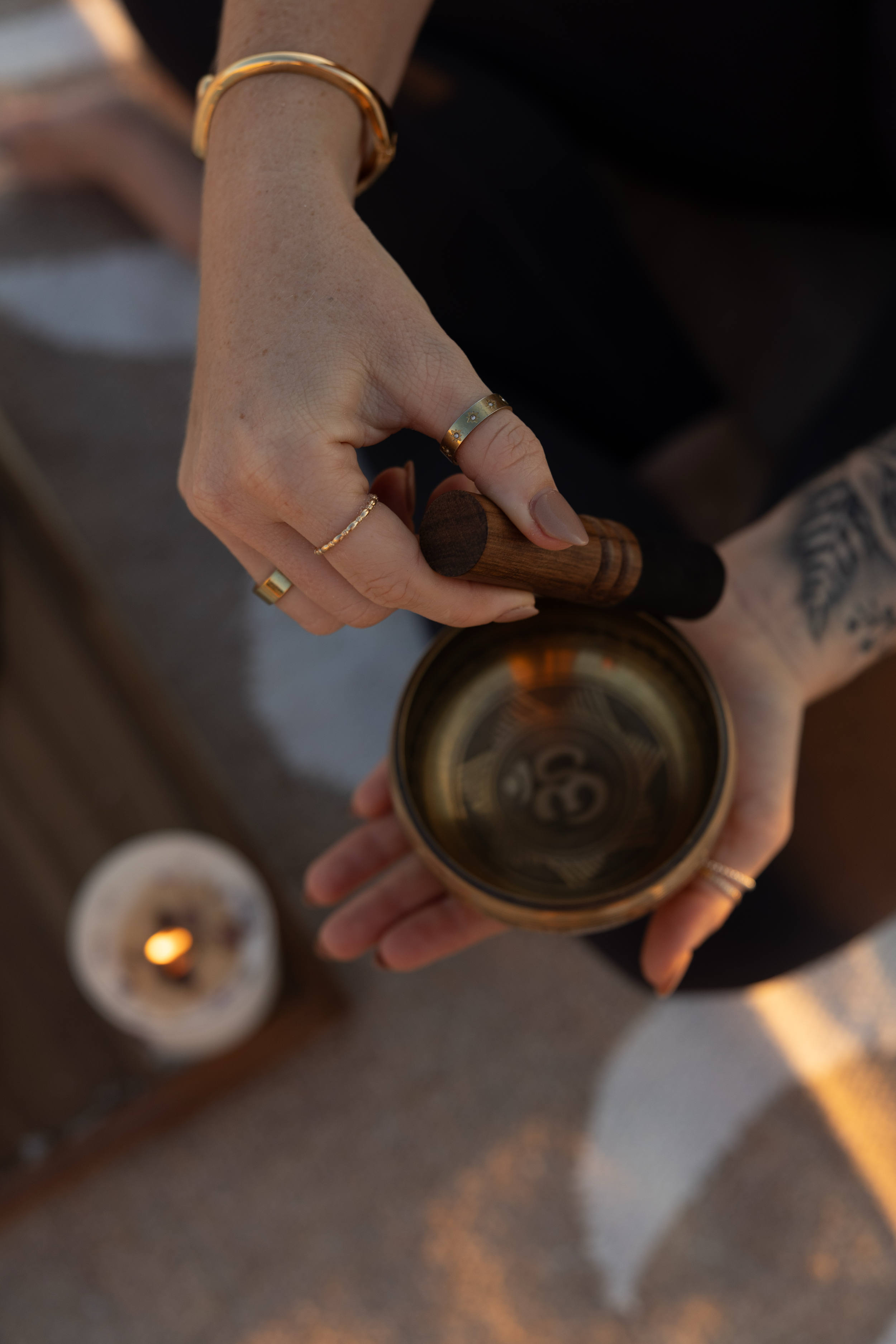Person holding a brass colored sound bowl with a wooden mallet, wearing multiple rings and a gold bracelet, with a lit candle in the background.