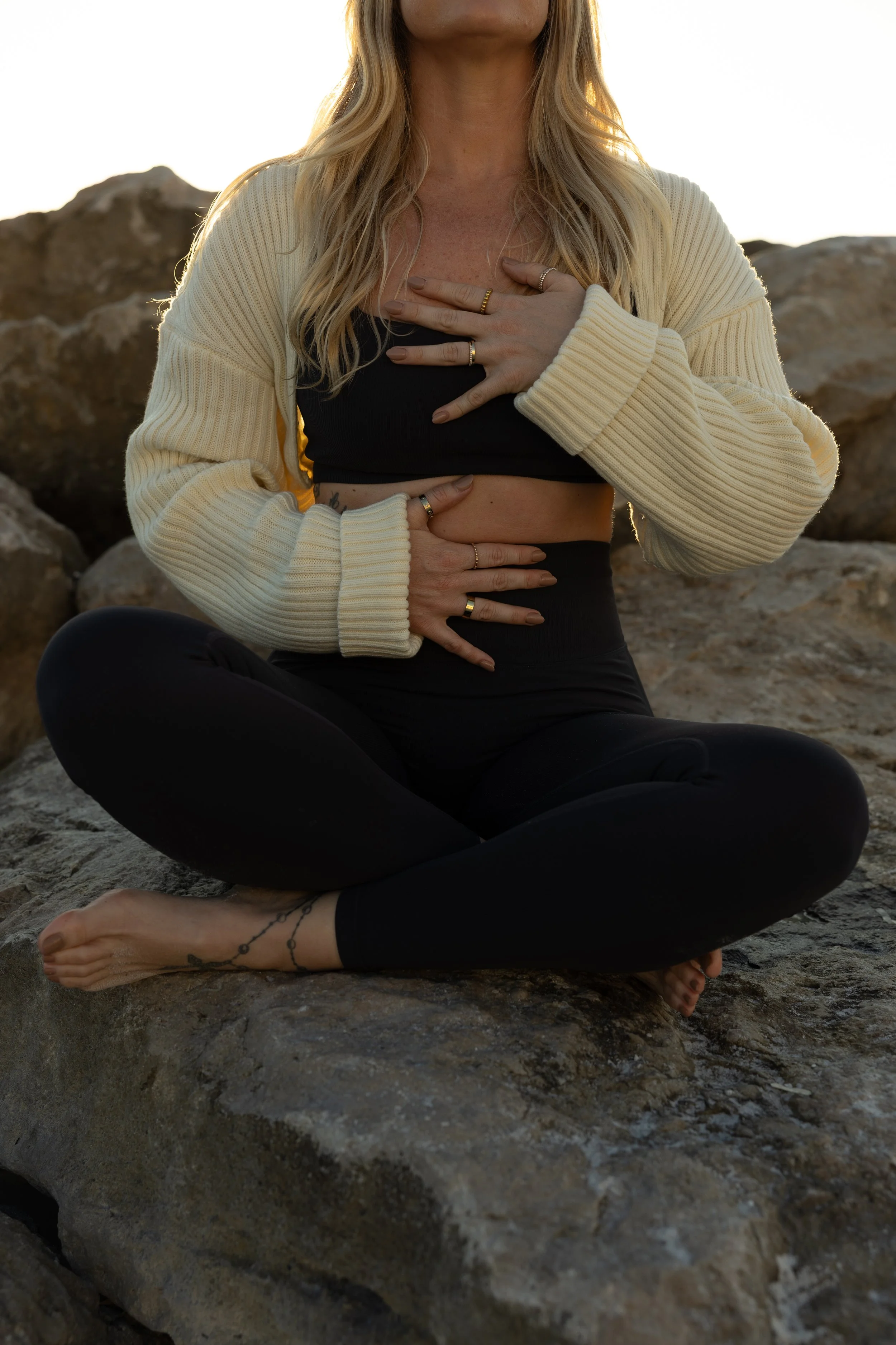 A woman sitting cross-legged on a rock, wearing black leggings, a black top, and a cream-colored cardigan, with her hands placed on her chest and stomach, palm down. She has long blonde hair and a tattoo on her left ankle. The background features rocky terrain and a bright sky.