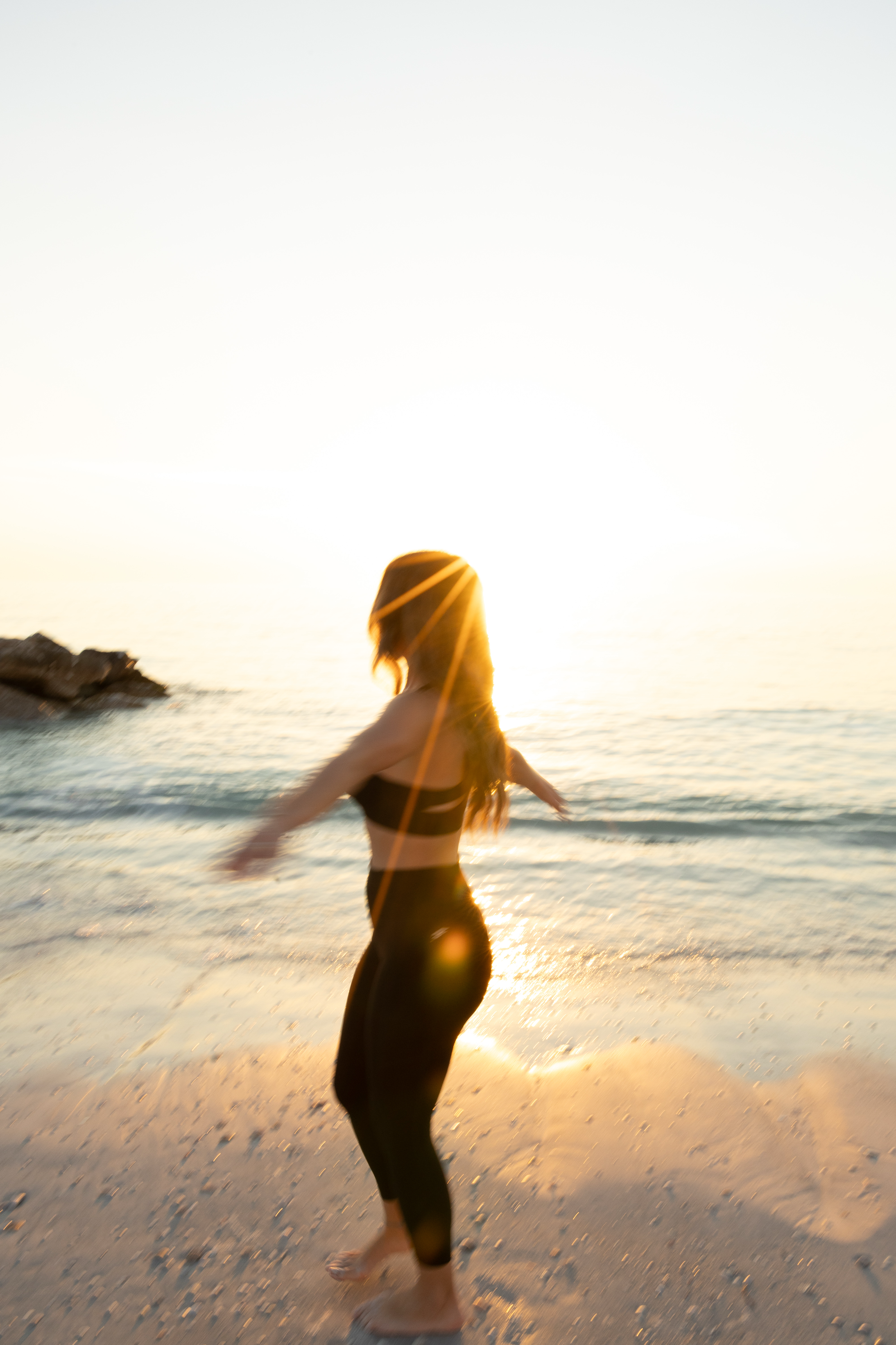 A woman dancing or playing on a beach at sunset with the ocean in the background.