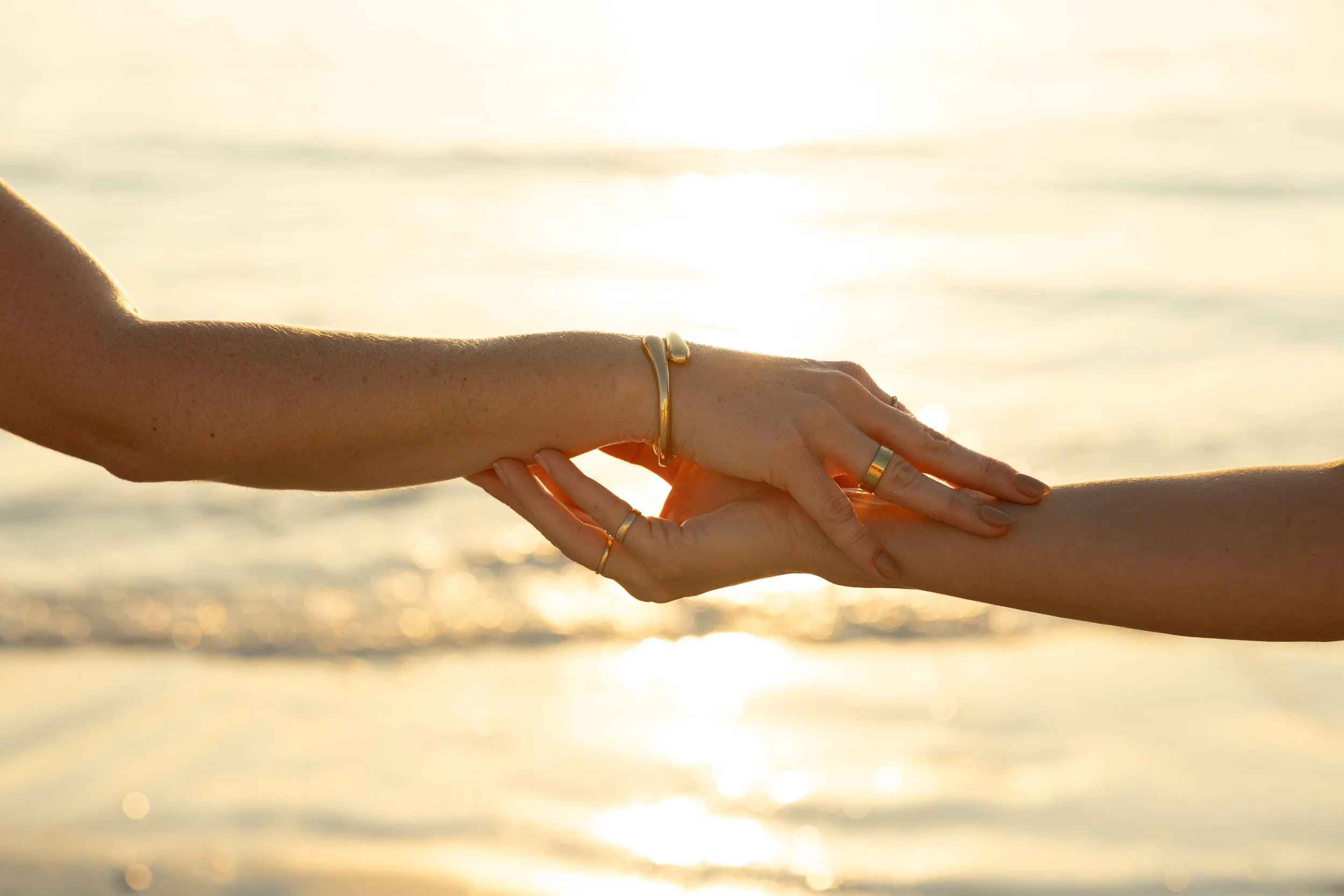 Two hands, one with a gold bracelet and rings, reaching out and holding each other against a sunset or sunrise over the water.