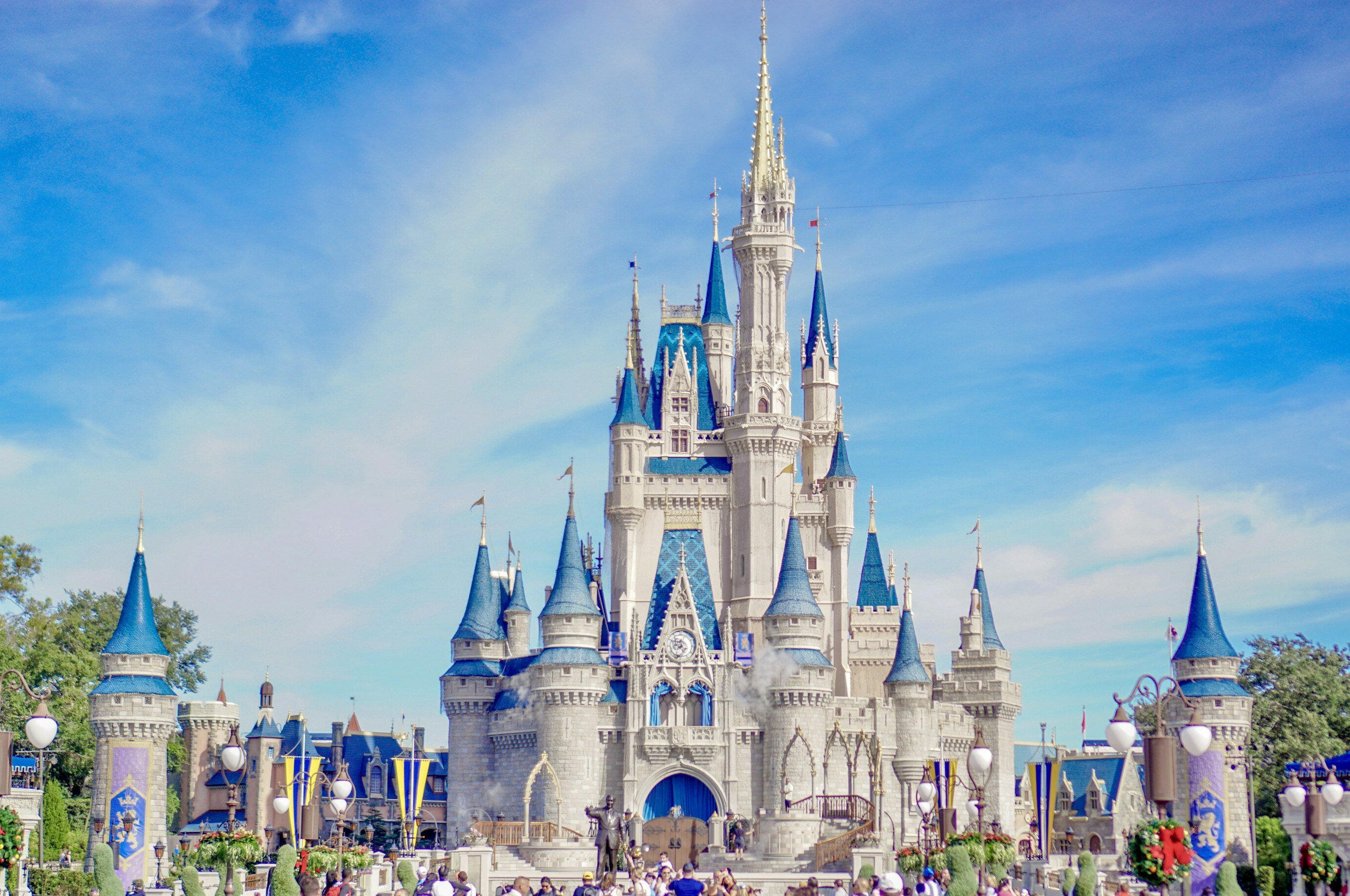 The Cinderella Castle at Disney World, a large fairy tale castle with blue rooftops, tall spires, and ornate details, set against a bright blue sky with some clouds.