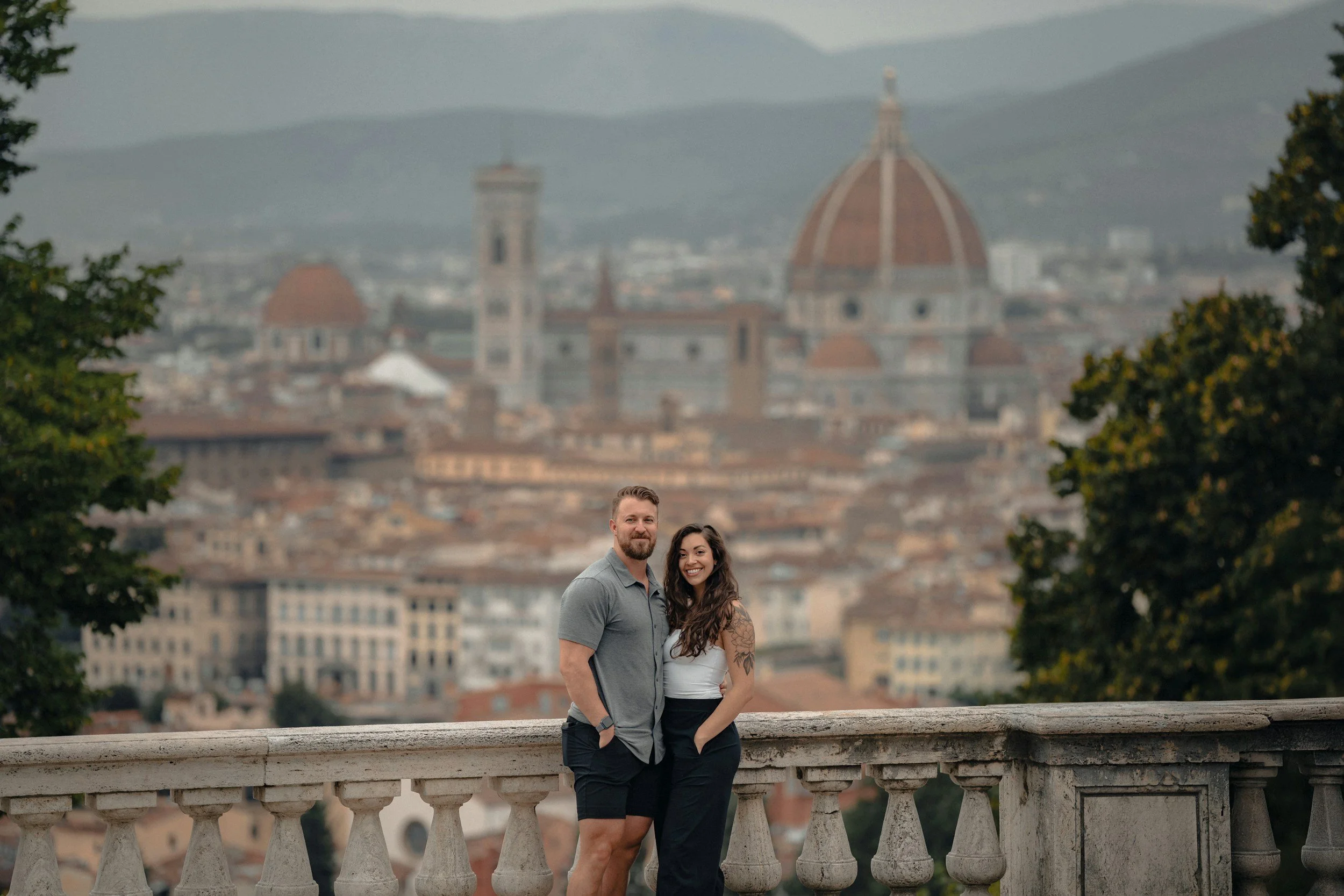 A man and woman standing close together on a stone balustrade overlooking the city of Florence, Italy, with the Florence Cathedral's dome in the background.