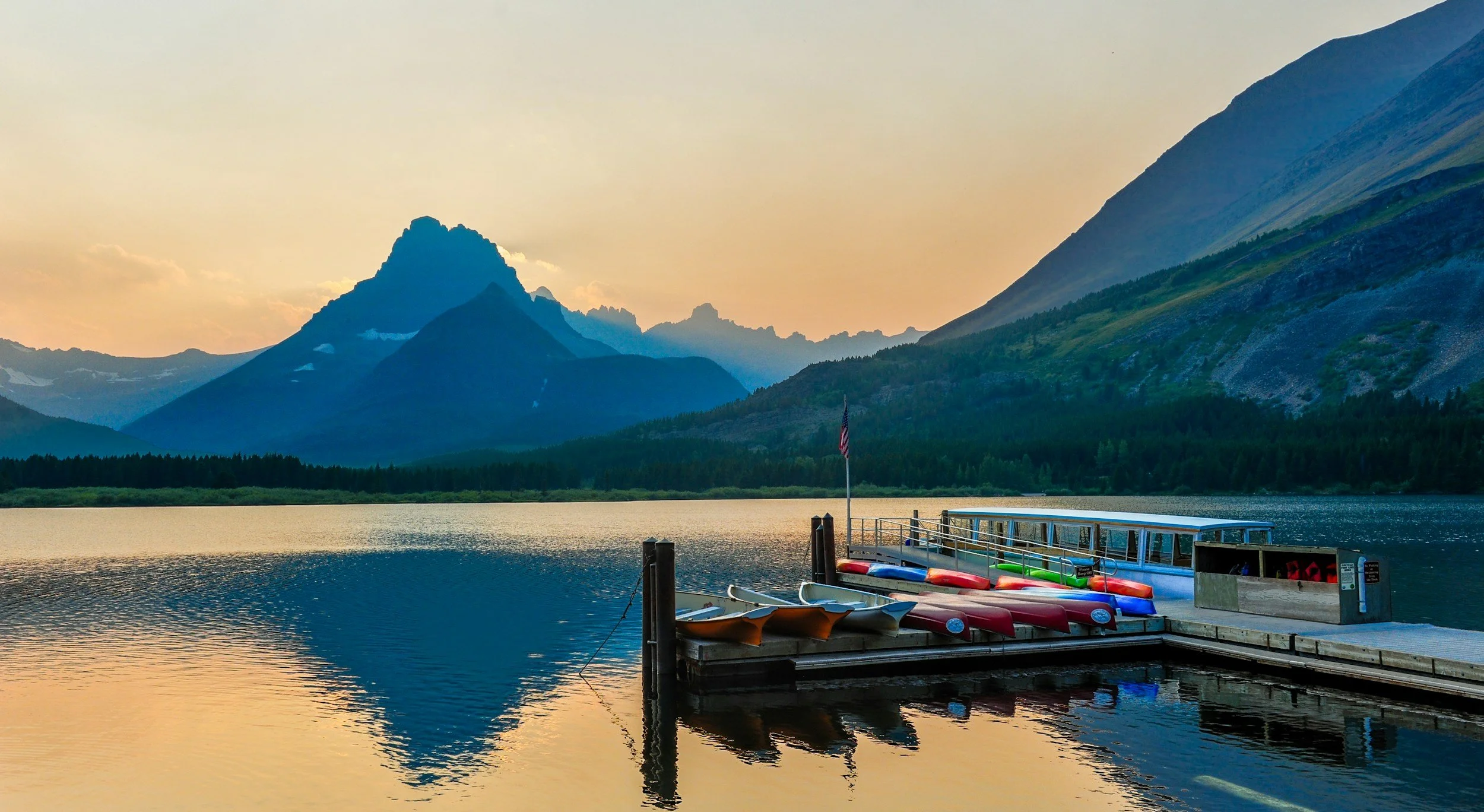 A dock with canoes and kayaks on a calm lake at sunset, surrounded by mountains and forest.