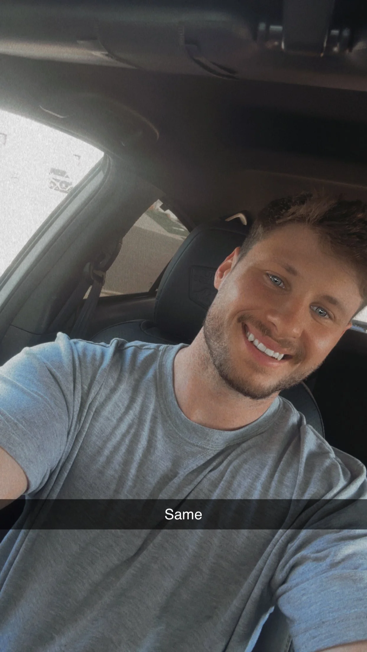 A young man with short brown hair, blue eyes, and a beard, smiling in a car. He is wearing a gray t-shirt, and the caption says "Same."