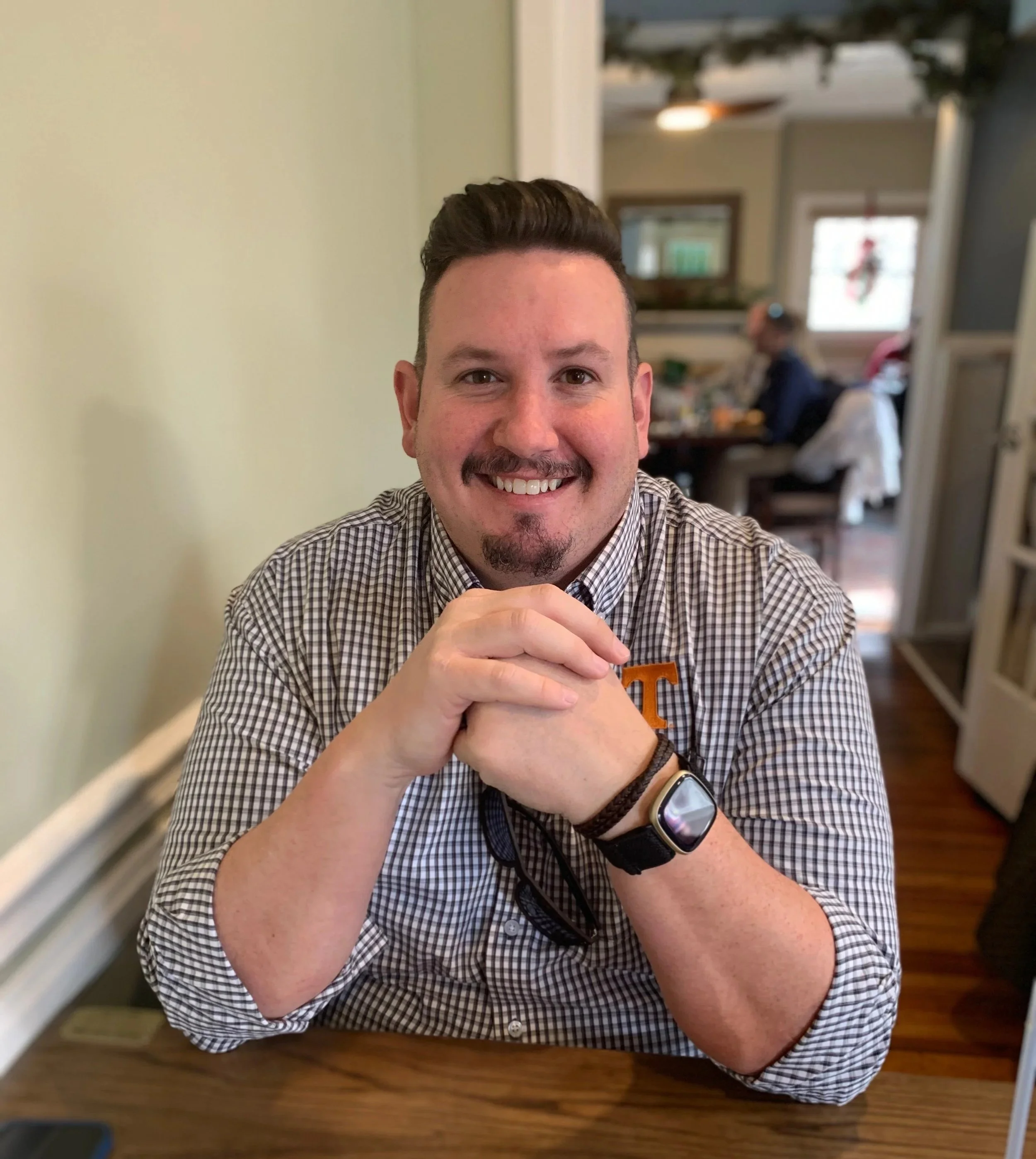 A smiling man with short dark hair and a beard, wearing a checkered shirt, sitting at a wooden table with his hands clasped, in a cozy, well-lit room with people in the background.