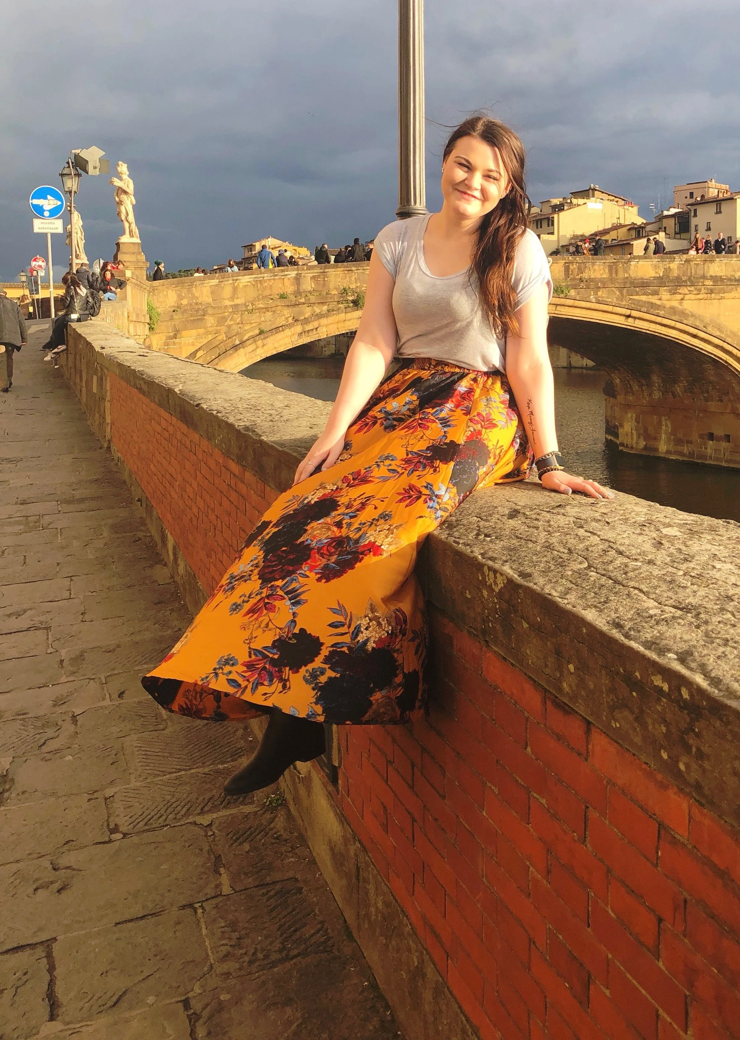 Young woman sitting on a brick bridge wall by a river, smiling, with dark clouds and historic statues and buildings in the background.