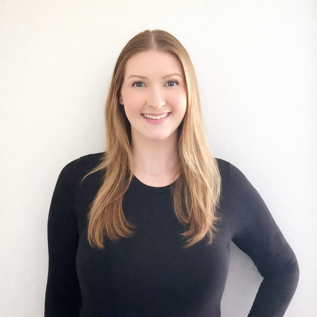 A young woman with long, light brown hair and blue eyes, smiling, wearing a black long-sleeve shirt, standing against a plain white wall.