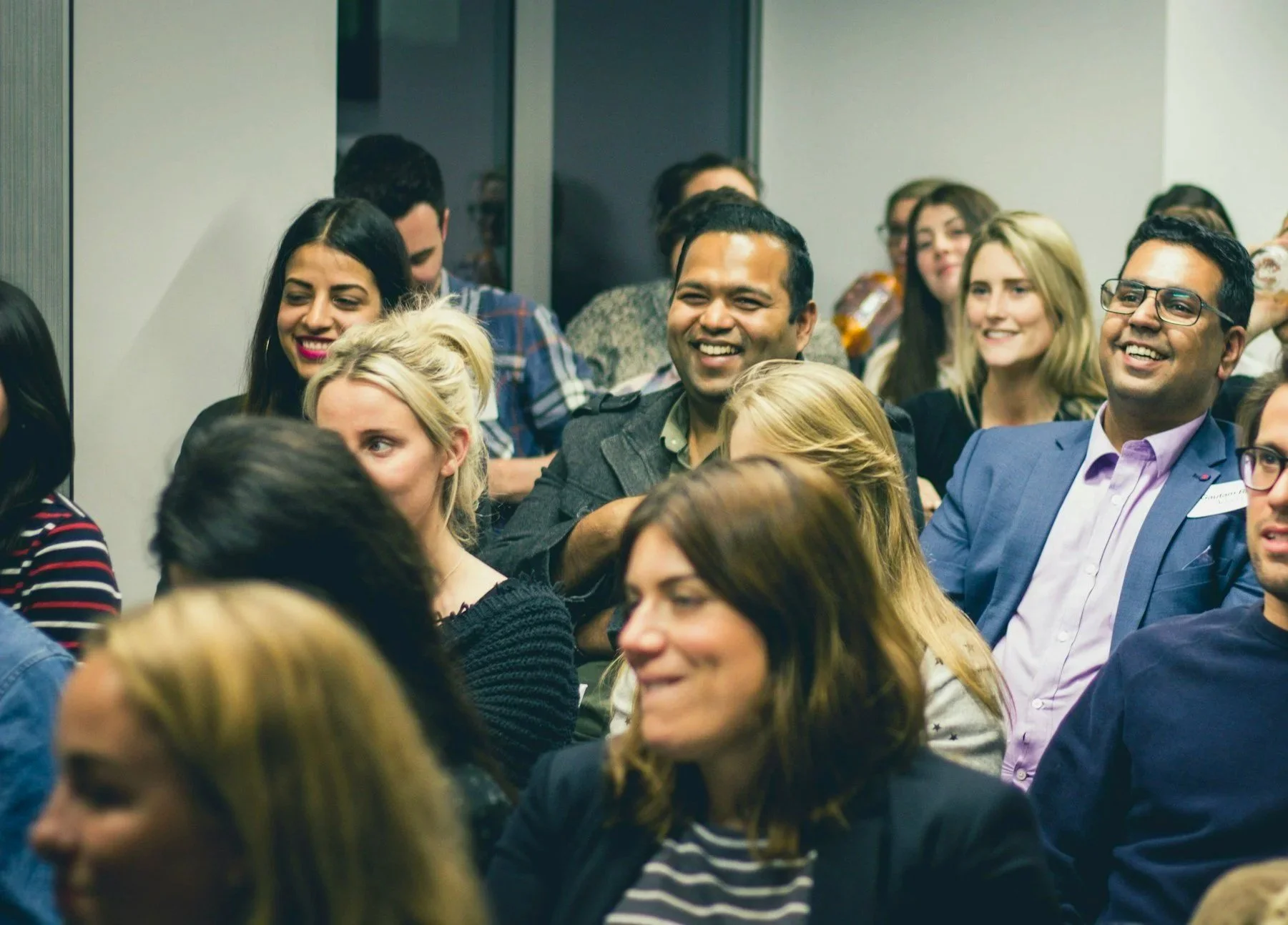 People attending a conference or seminar, sitting and smiling in a well-lit indoor setting.