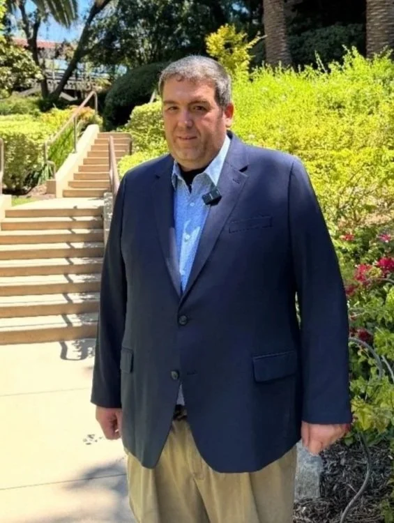 A man in a navy blazer and beige pants standing outside on a sunny day with stairs and greenery in the background.