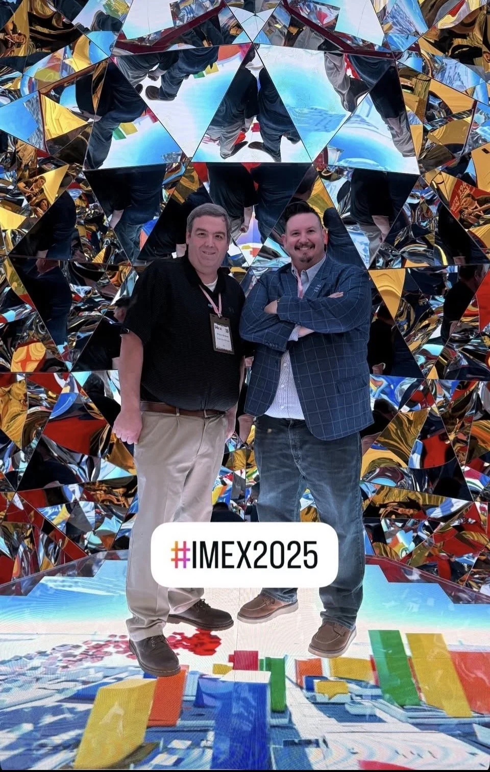 Two men standing in front of a mirrored and colorful abstract installation at IMEX 2025 conference, with confetti and reflections visible.