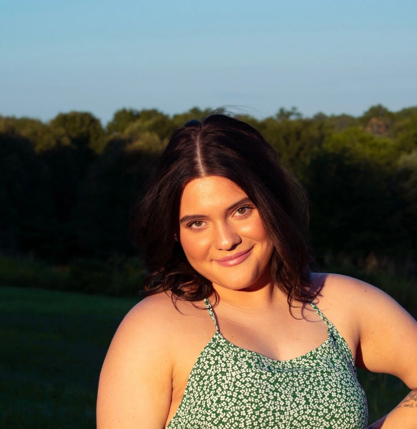 A woman with dark brown hair in a green floral dress outdoors at sunset, smiling at the camera with a forest background.