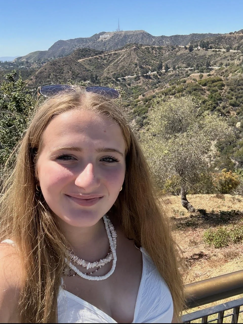 A young woman with long blonde hair wearing a white top and shell necklace, taking a selfie outdoors with a mountainous landscape and the Hollywood sign in the background.