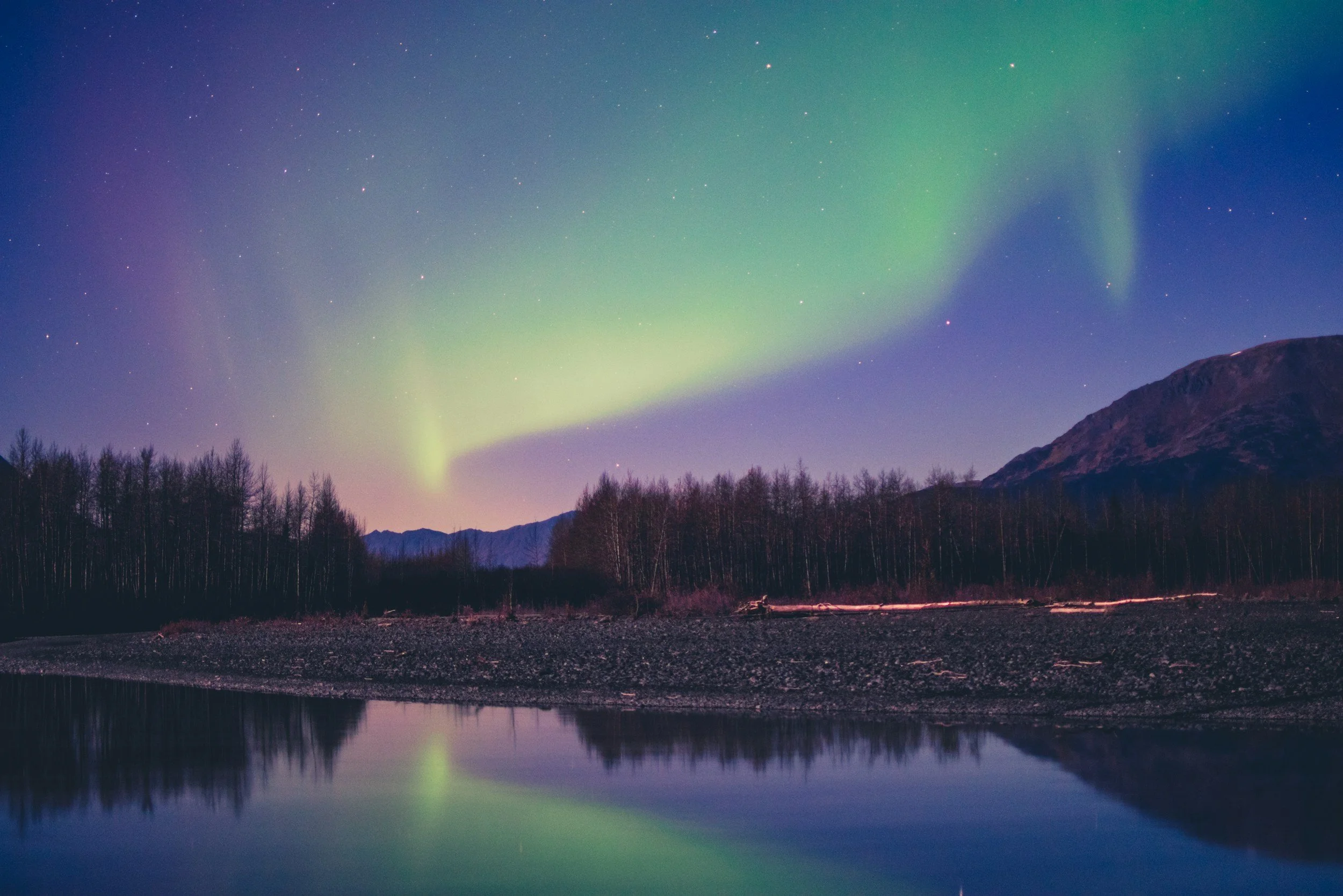 Northern lights over a river, with a forest and mountains in the background at dusk or night.