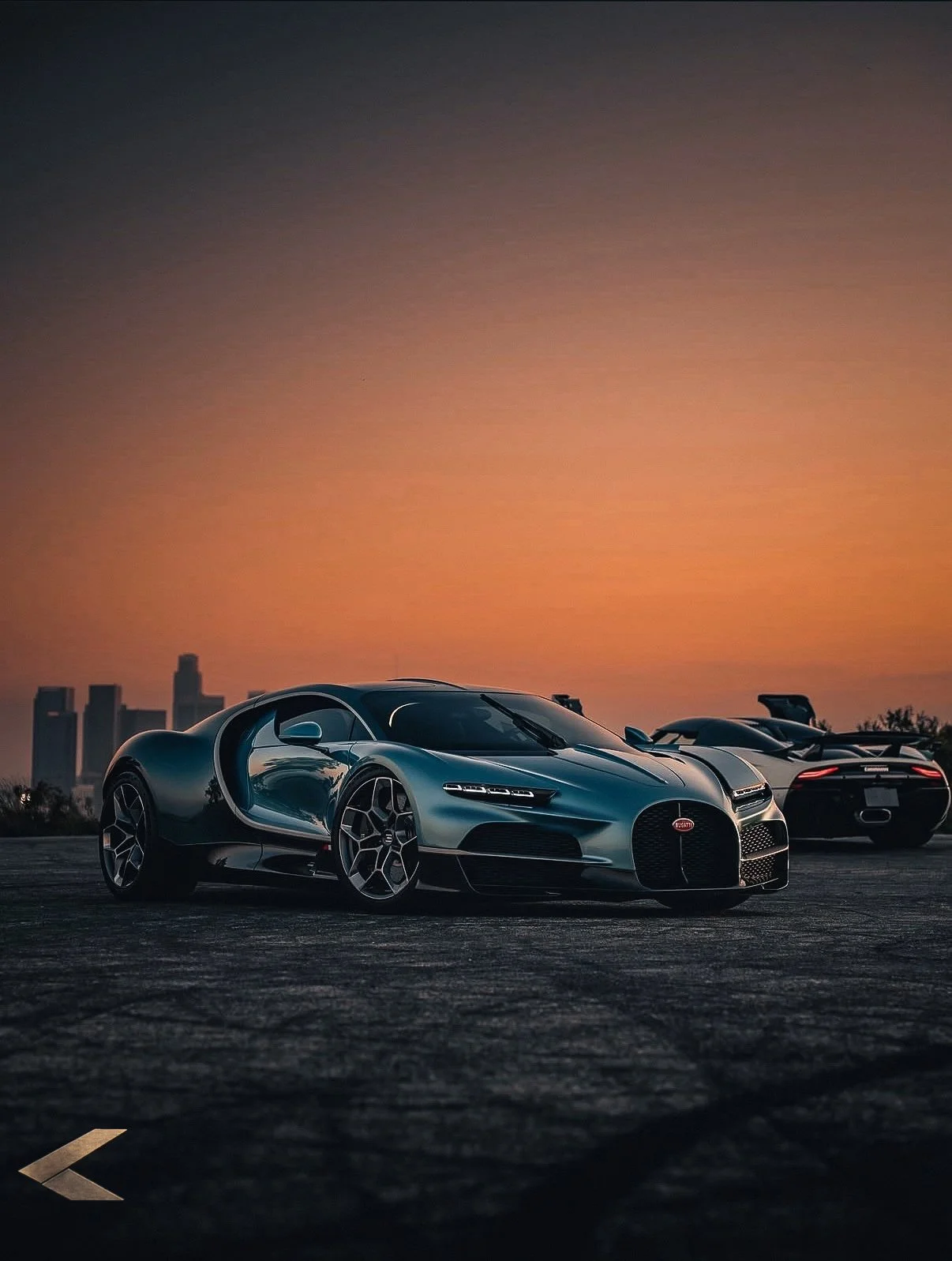 Two luxury sports cars parked on an open road at sunset with city skyline in the background. The car in front is a silver Bugatti with a sleek design, and behind it is a black sports car. The sky has a warm orange hue, creating a dramatic backdrop.
