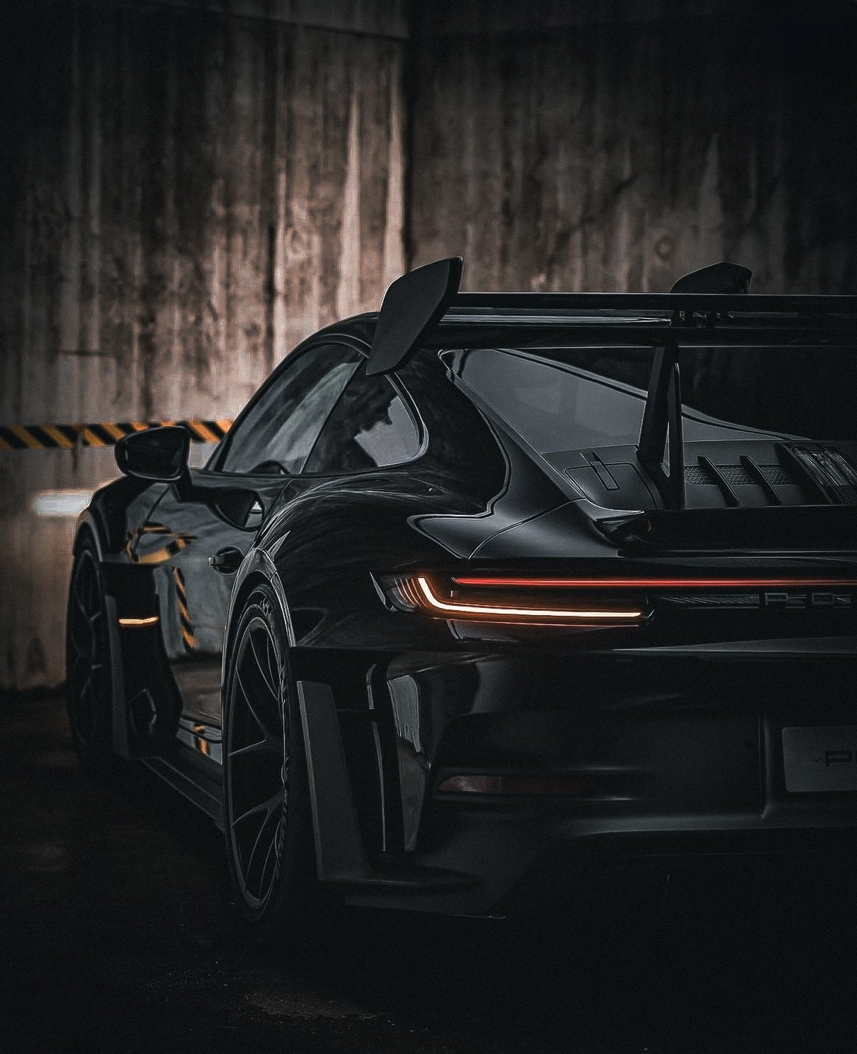 Black Porsche sports car with rear wing, illuminated tail lights, parked in a dimly lit garage with wooden wall background.