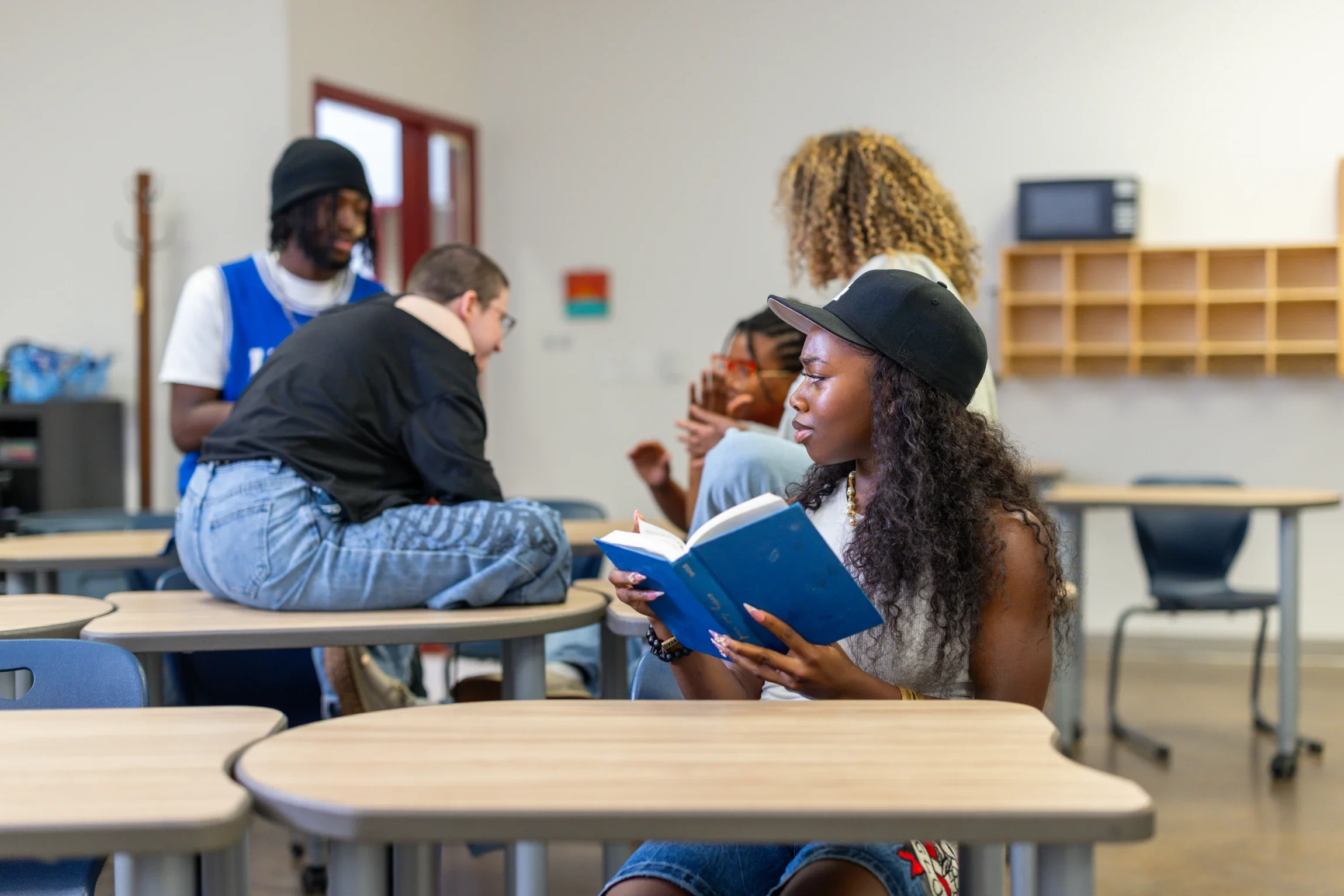A high school student holding a blue book. She is looking up from her book as she listens in on other teenagers in the background.
