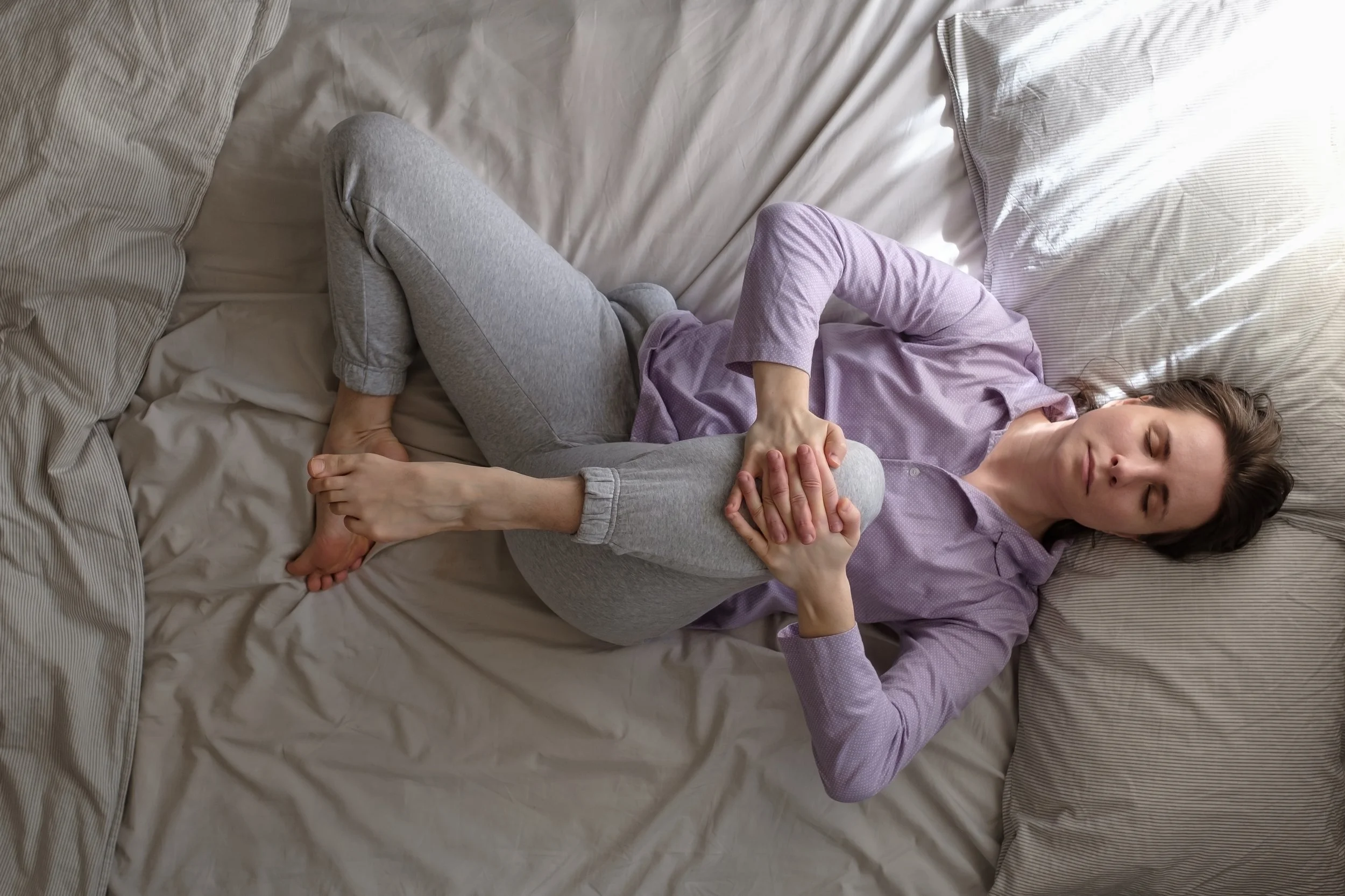 Top-down view of a woman in purple pajamas stretching her hips and knees in bed, seeking relief from pelvic floor tension at Signature PT in Manhattan.