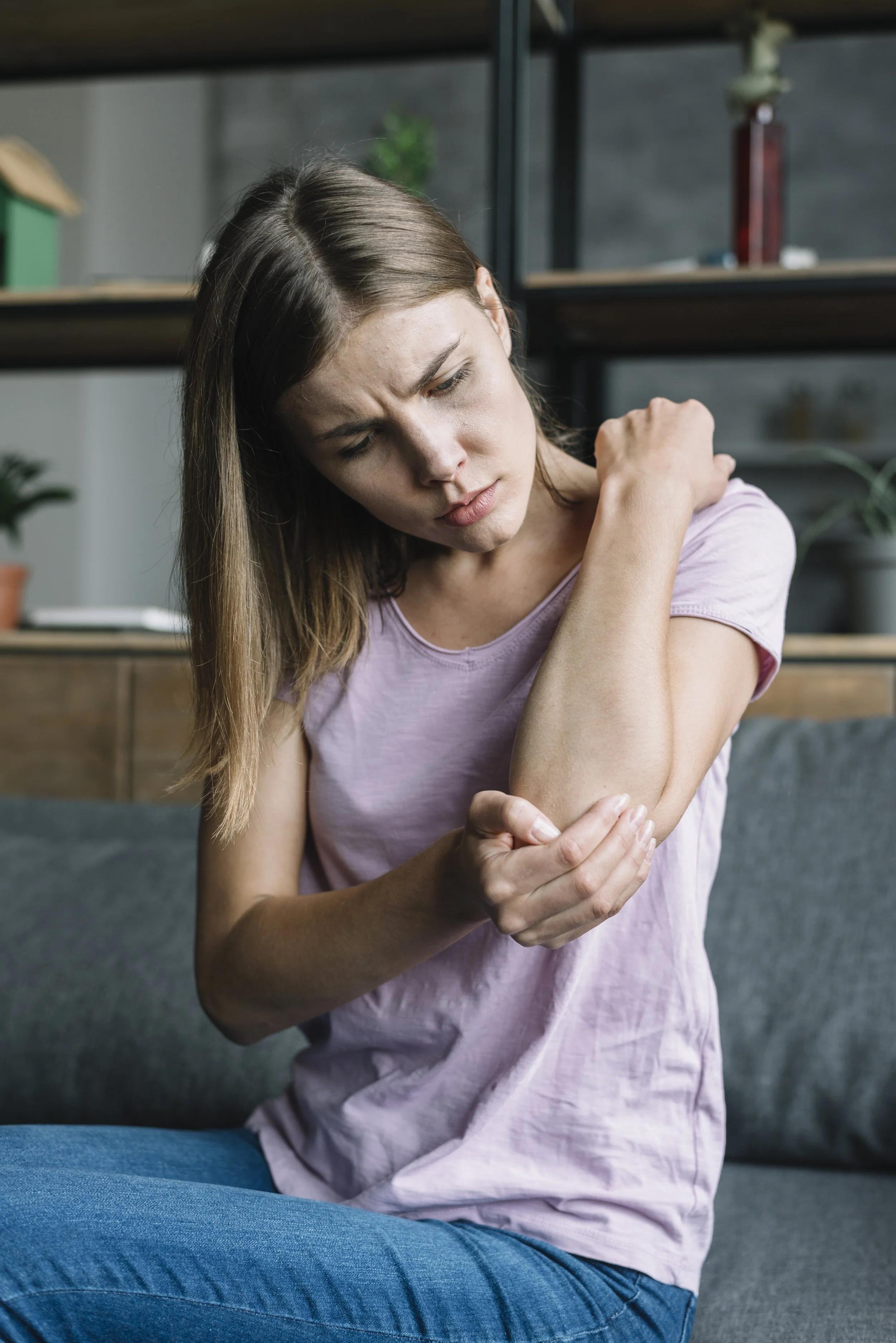 A woman sitting on a sofa examining her painful elbow, illustrating symptoms of repetitive strain, tendonitis, and joint stiffness.