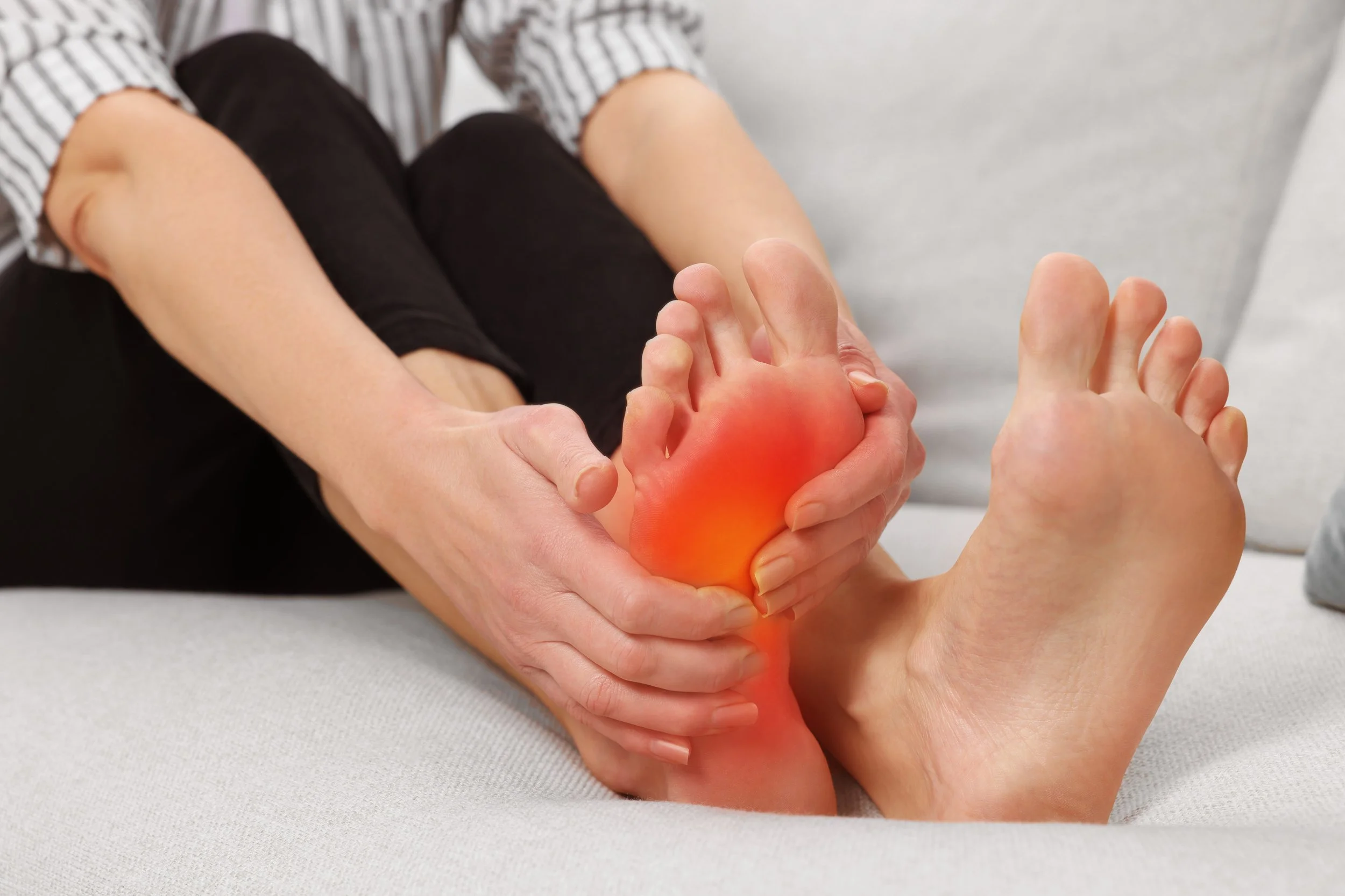 Close-up of a person on a sofa clutching a painful foot with a red pain indicator, seeking foot and ankle physical therapy at Signature PT in Manhattan.