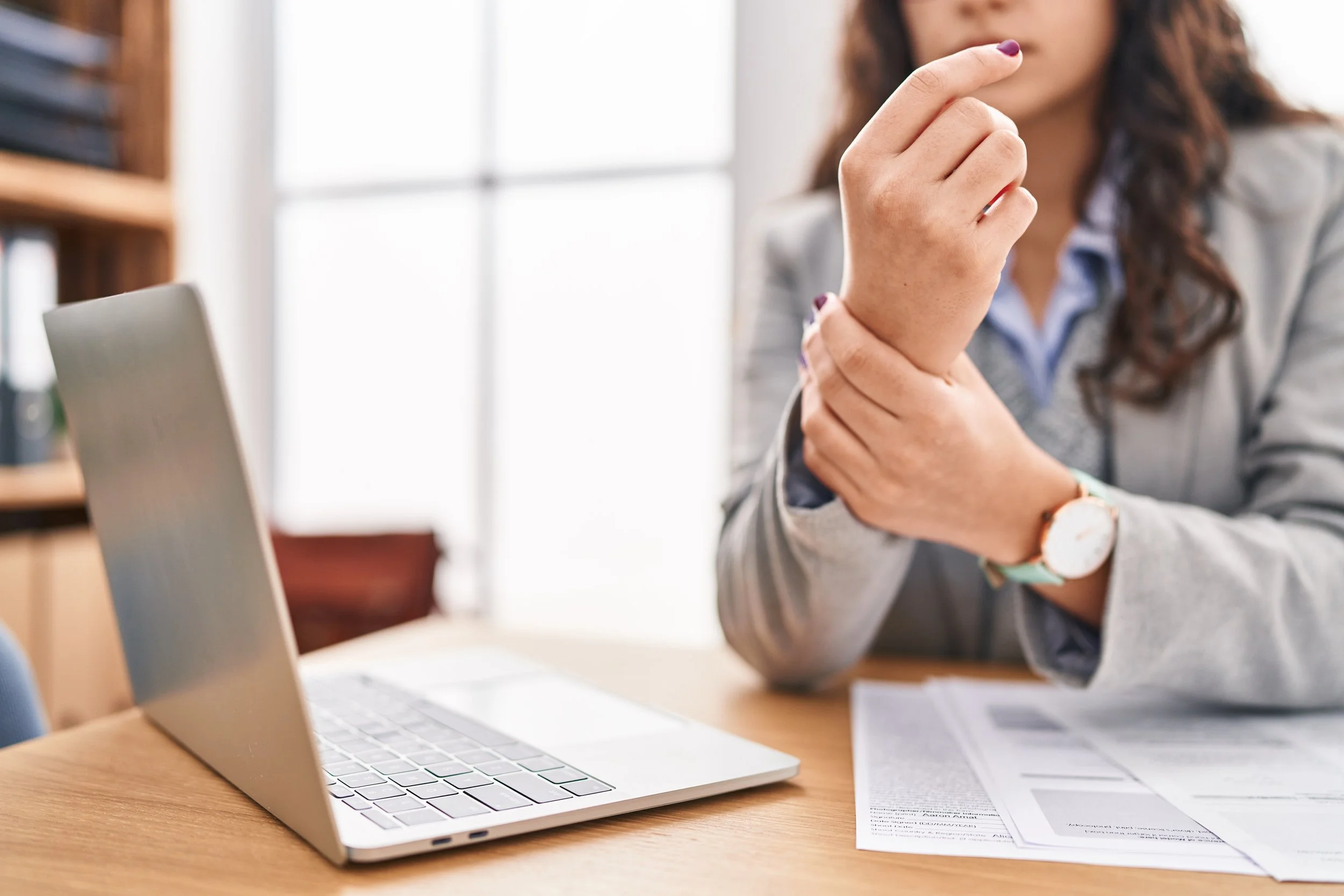 A woman in a grey blazer sitting at a desk and clutching her painful wrist, seeking carpal tunnel and repetitive strain relief at Signature PT in Manhattan.
