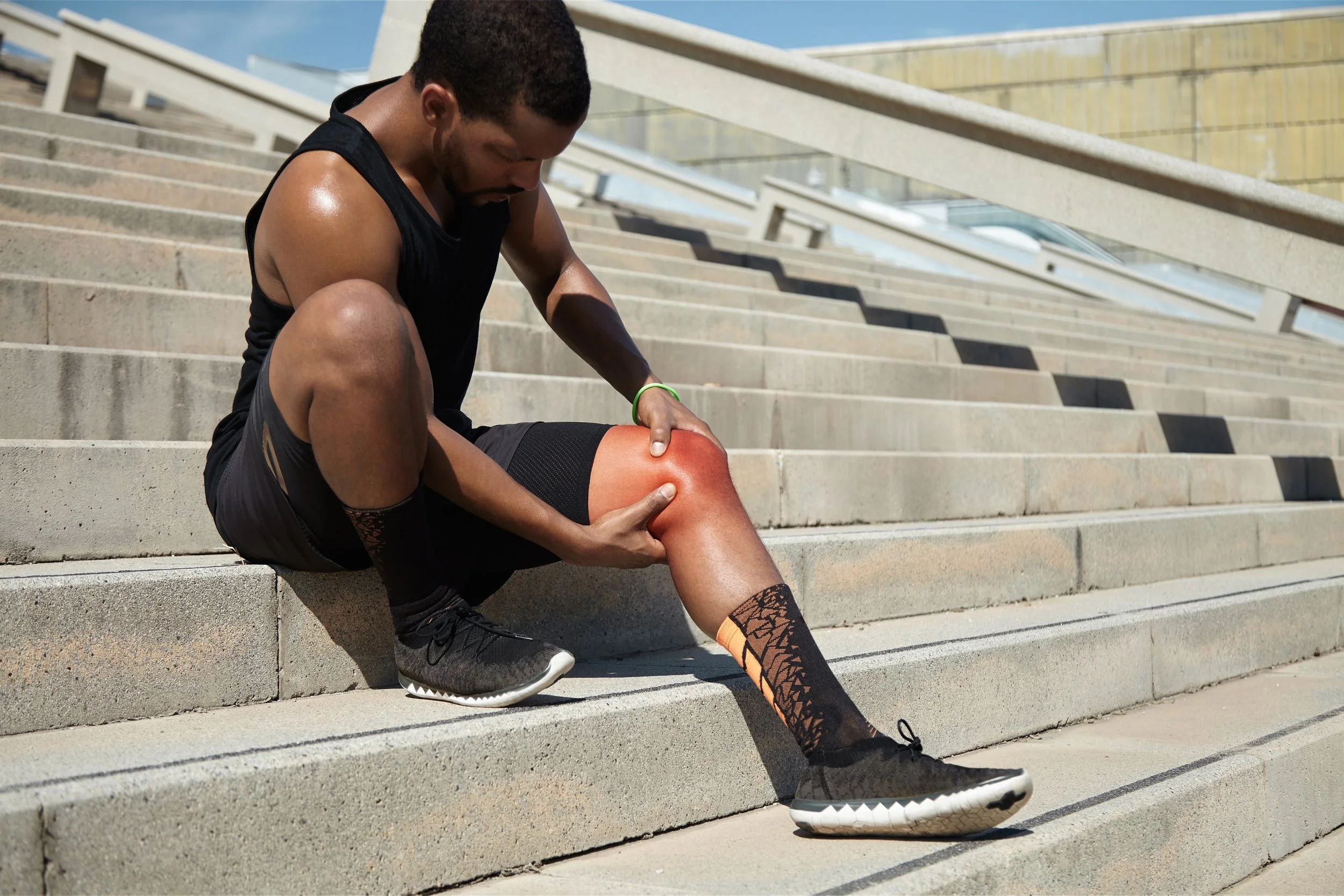 A male athlete sitting on concrete city stairs, clutching a painful knee with a red pain indicator, seeking sports injury rehabilitation in Manhattan.