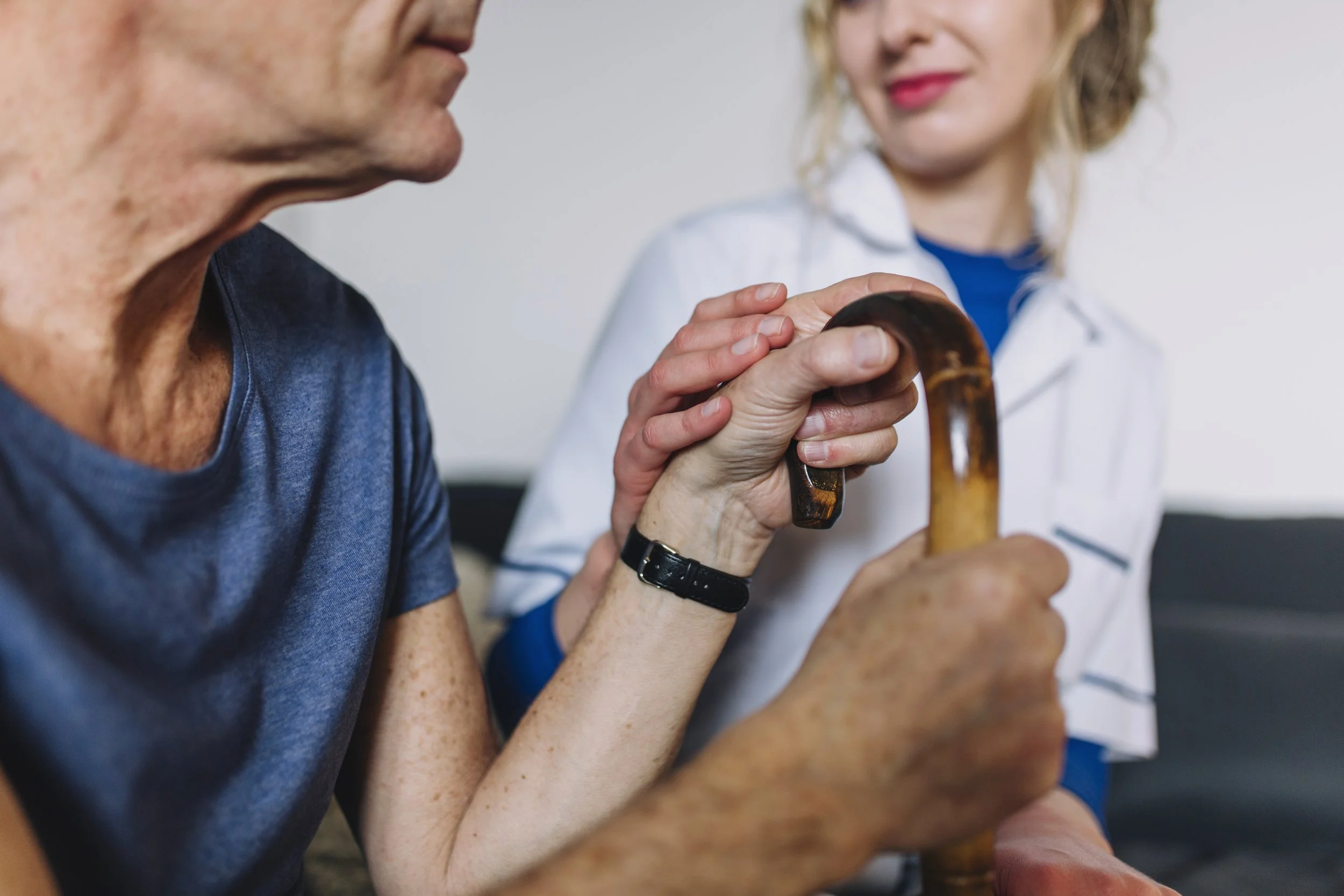 Close-up of a stroke patient's hand on a wooden cane being supported by a therapist, illustrating the balance and mobility challenges addressed in neuro-rehabilitation.
