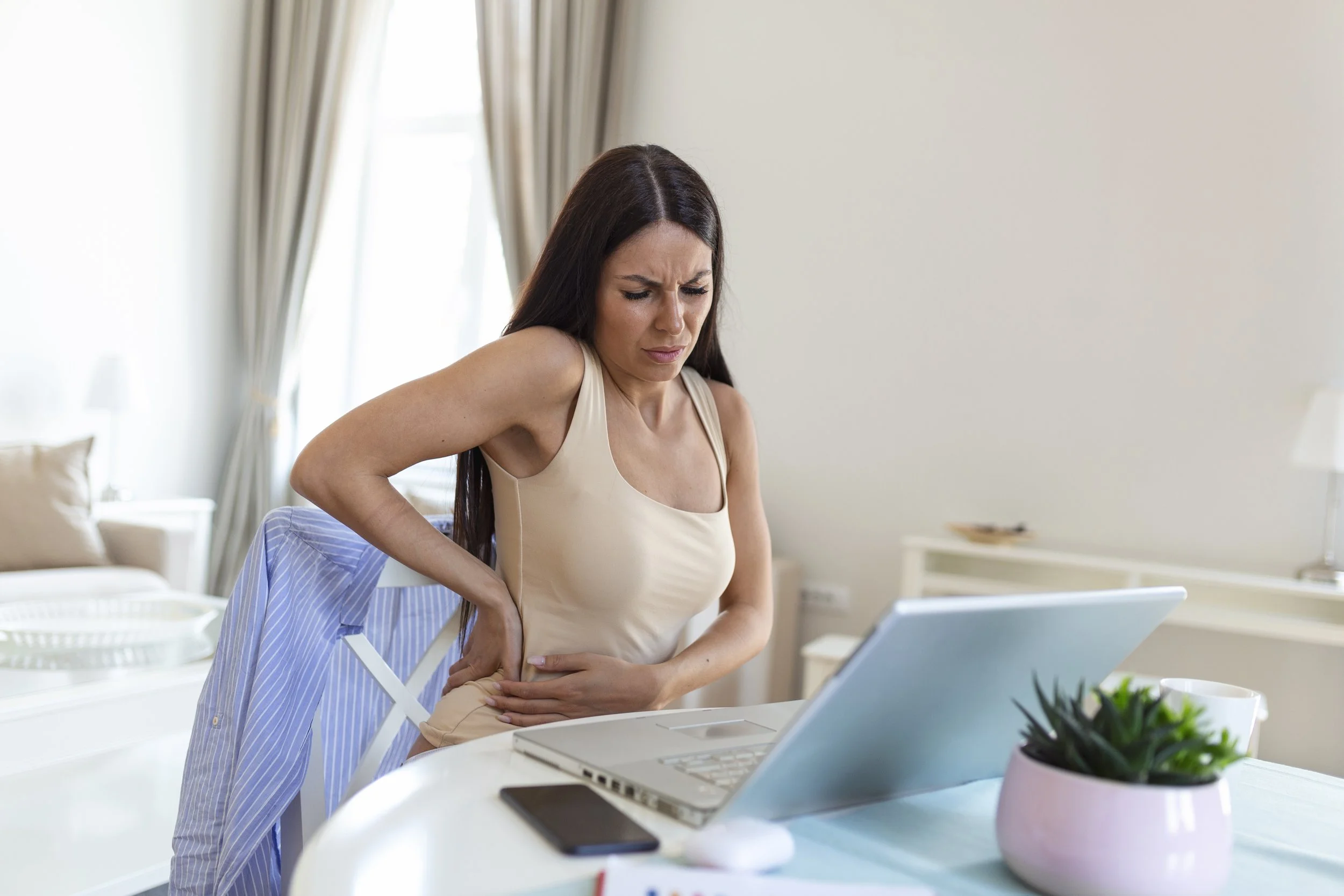 A professional woman in a home office clutching her painful lower back and hip, seeking relief from sitting-related hip pain at Signature PT in Manhattan.