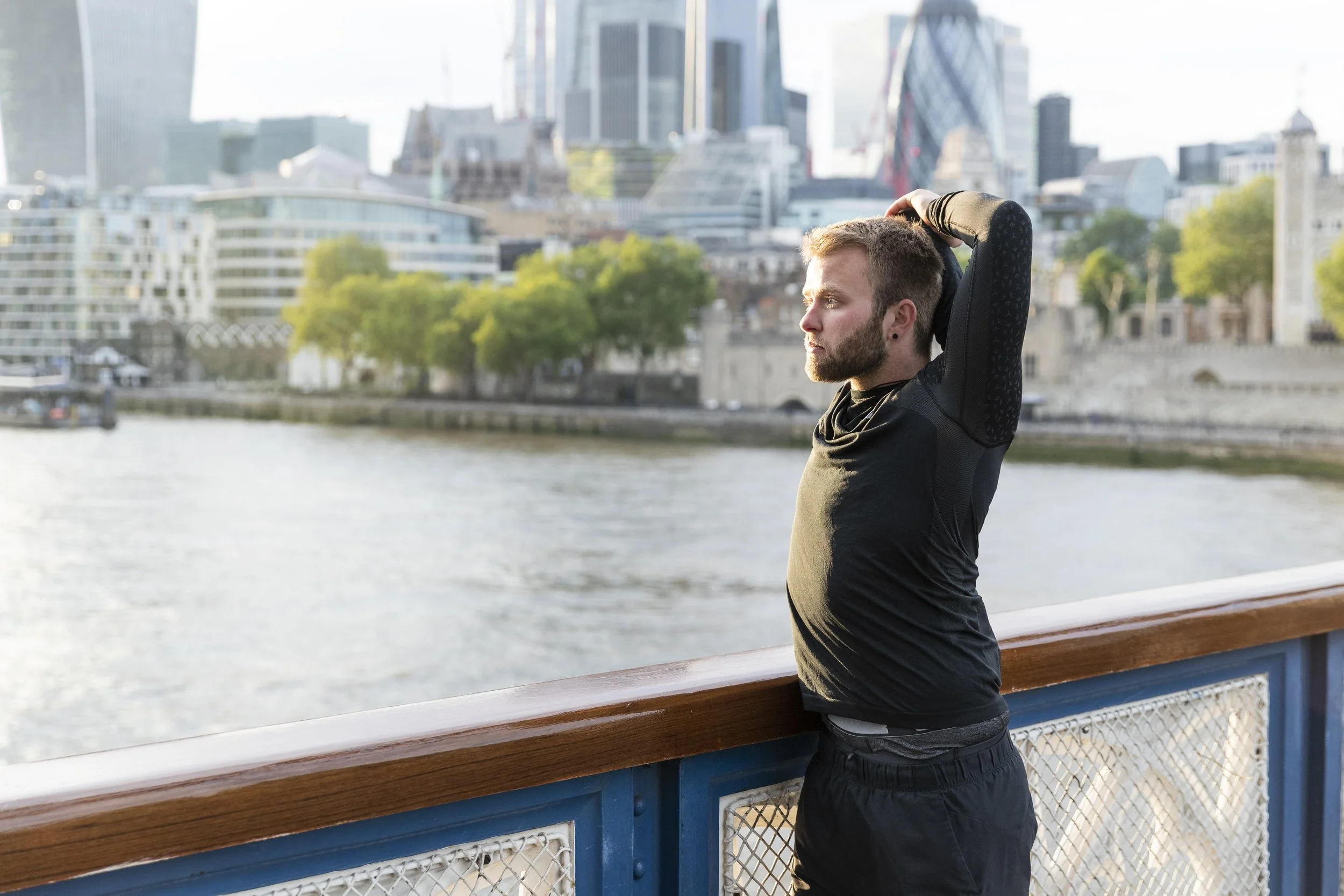A man stretching his arm and shoulder outdoors with a city skyline and river in the background, representing active recovery and physical therapy results.