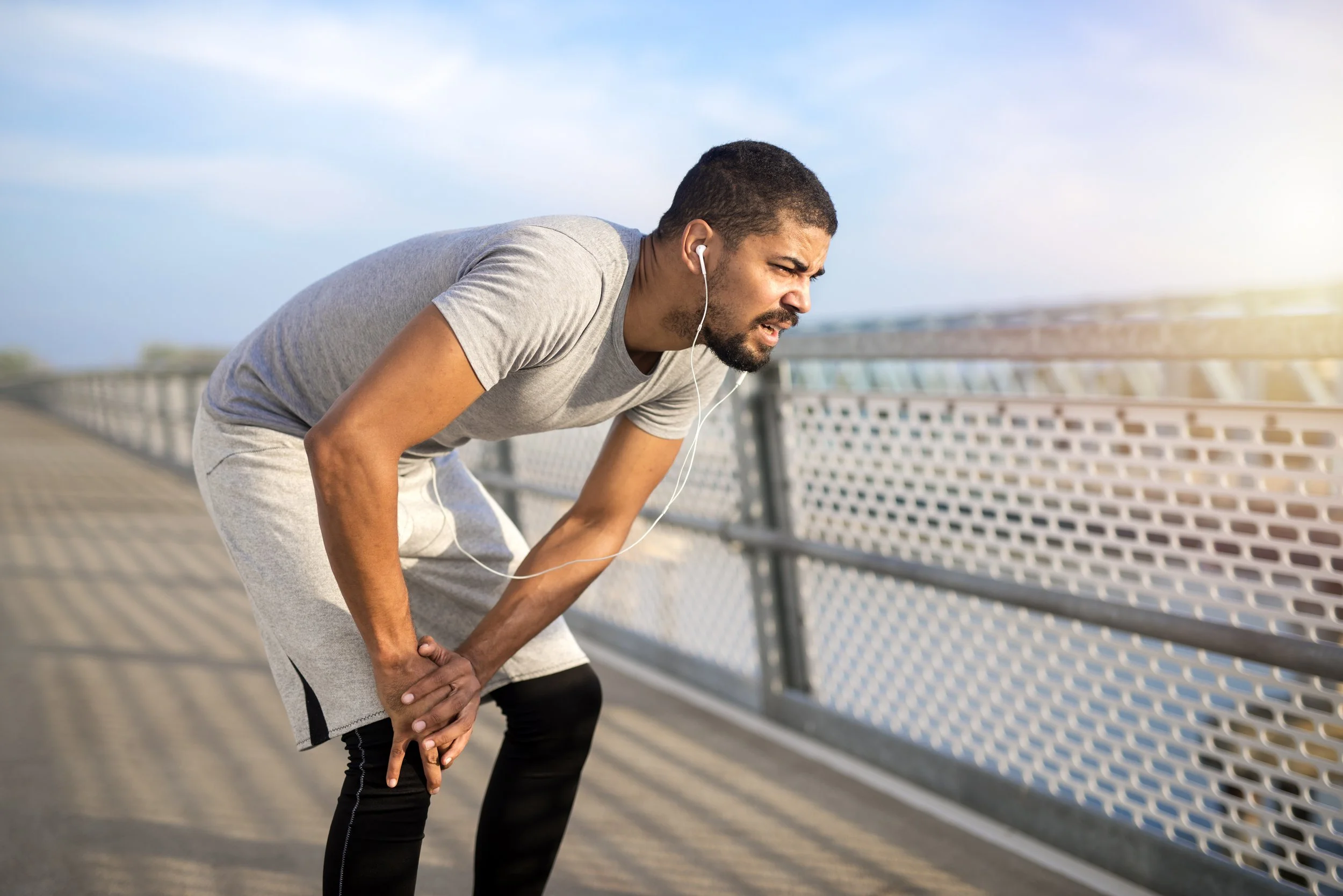 A male runner holding his injured knee while working out on a bridge in Manhattan, seeking sports medicine physical therapy at Signature PT.