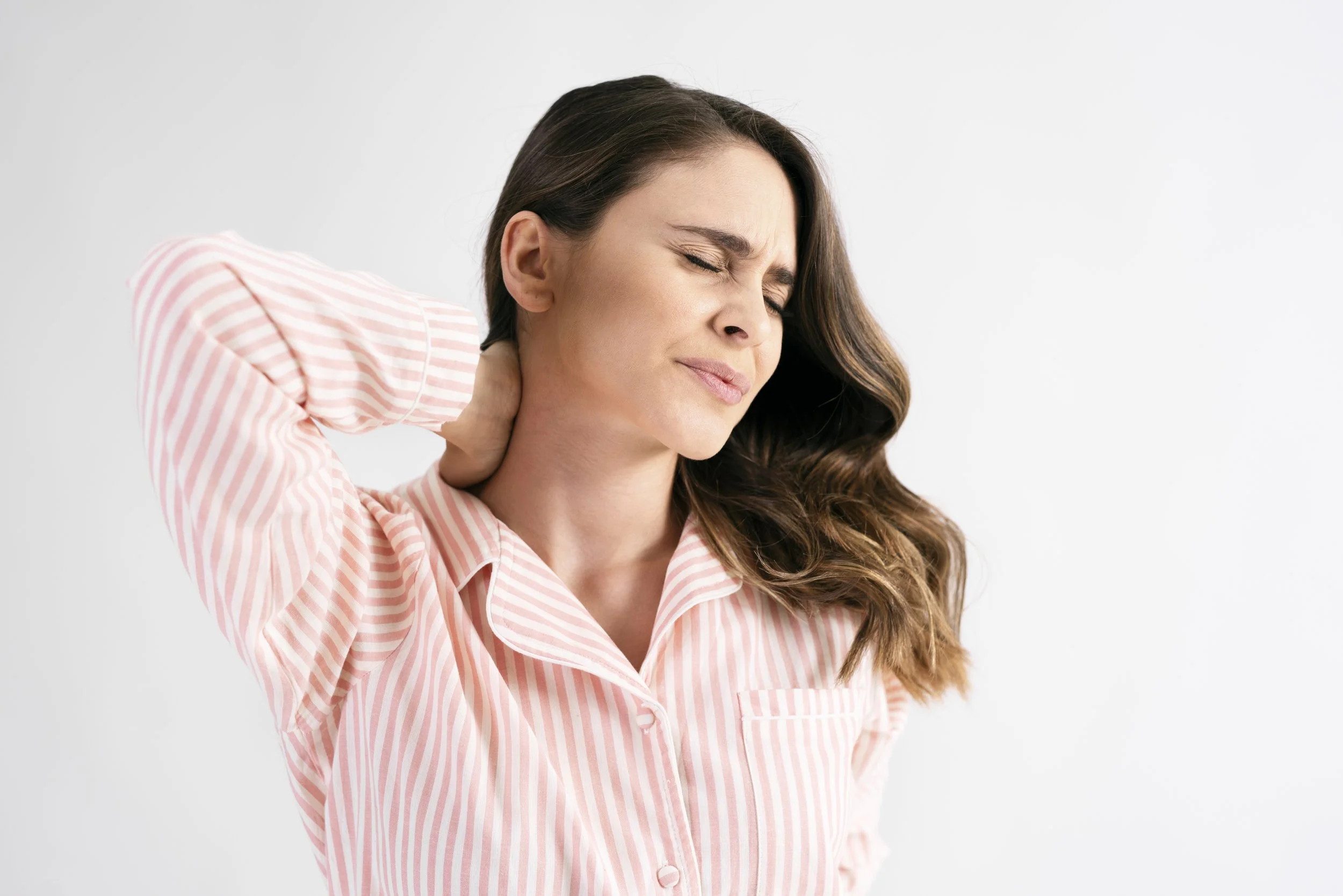 A woman in pink striped pajamas clutching her stiff and painful neck, seeking physical therapy for cervical relief at Signature PT in Manhattan.