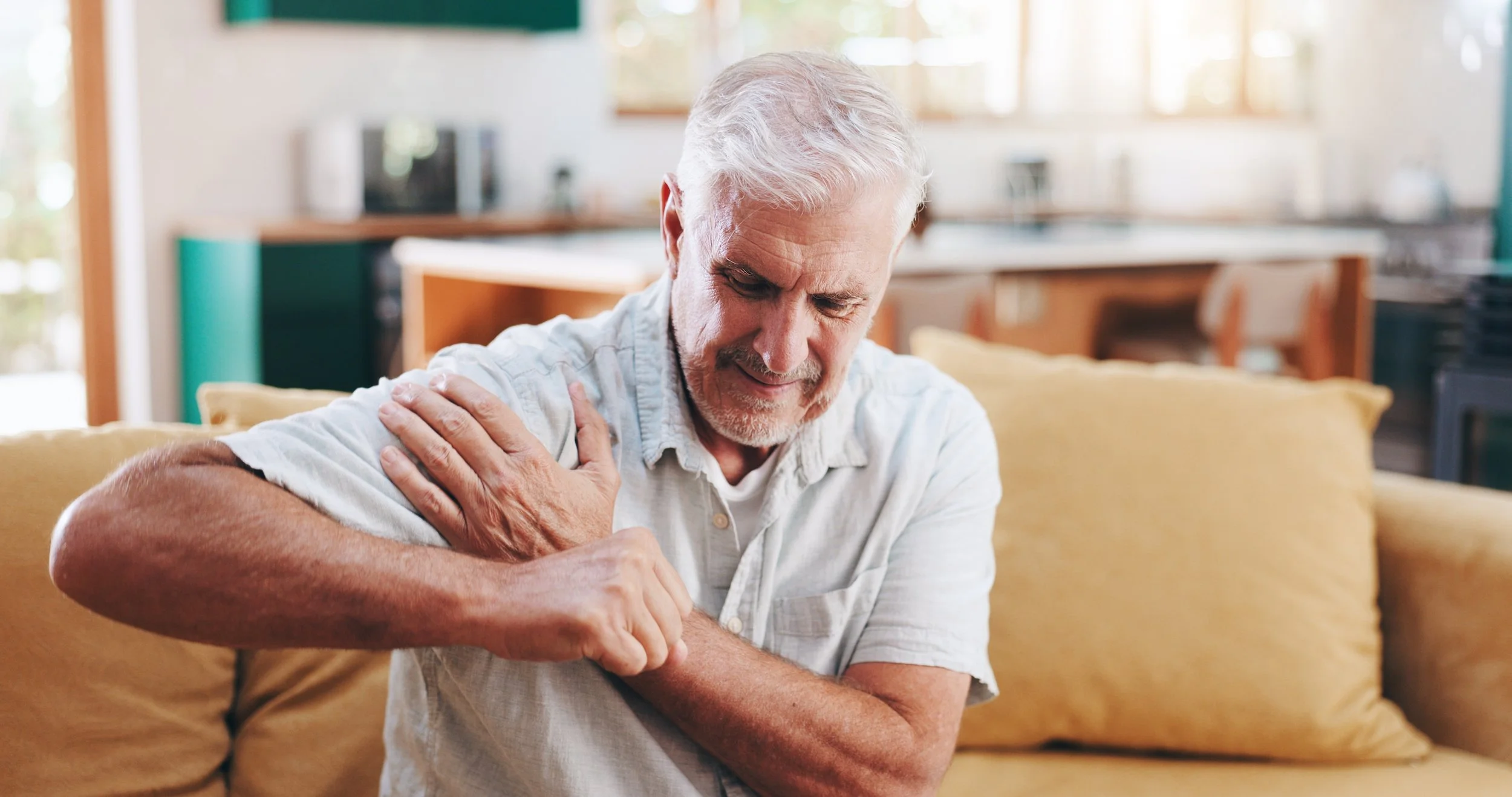 An older man sitting on a sofa clutching a painful shoulder, seeking relief from chronic joint pain at Signature PT in Manhattan.