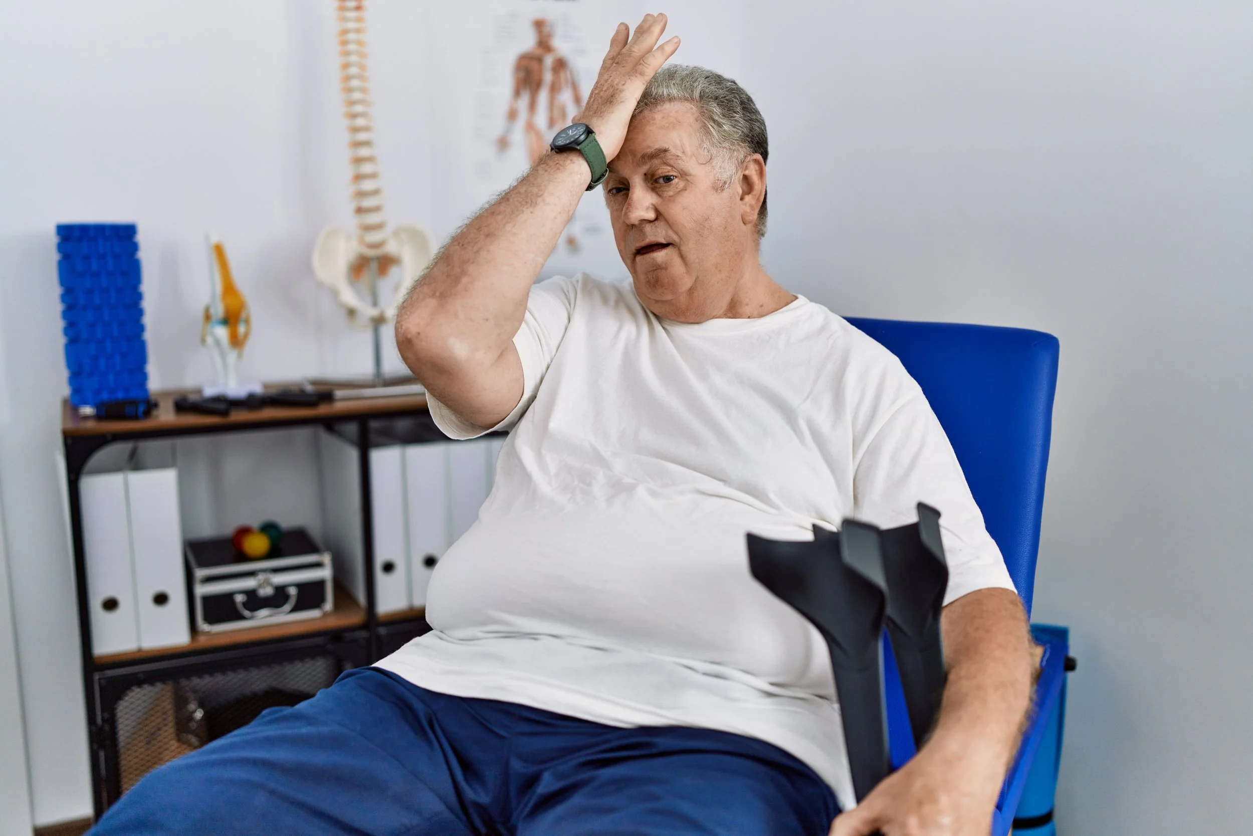 An older man sitting in a Manhattan physical therapy clinic with crutches, holding his forehead in frustration from neurological mobility challenges.