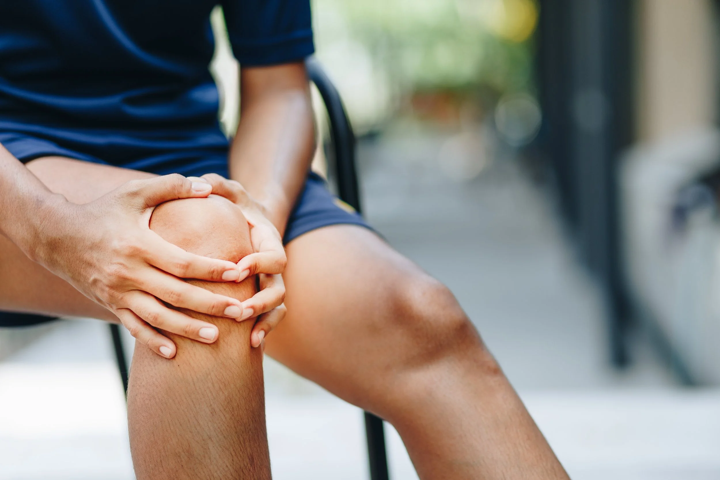 A person wearing athletic shorts clutches an injured, painful knee with a red pain indicator, seeking joint pain and sports medicine physical therapy at Signature PT in Manhattan.