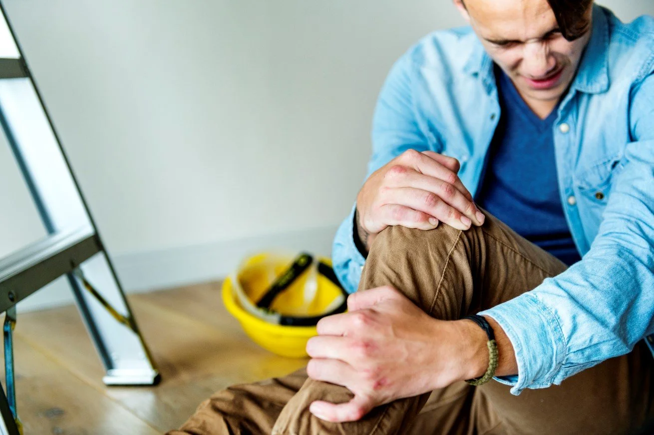 A worker holding an injured knee near a yellow hard hat and a ladder, seeking a worker's compensation physical therapy evaluation in Manhattan at Signature PT.